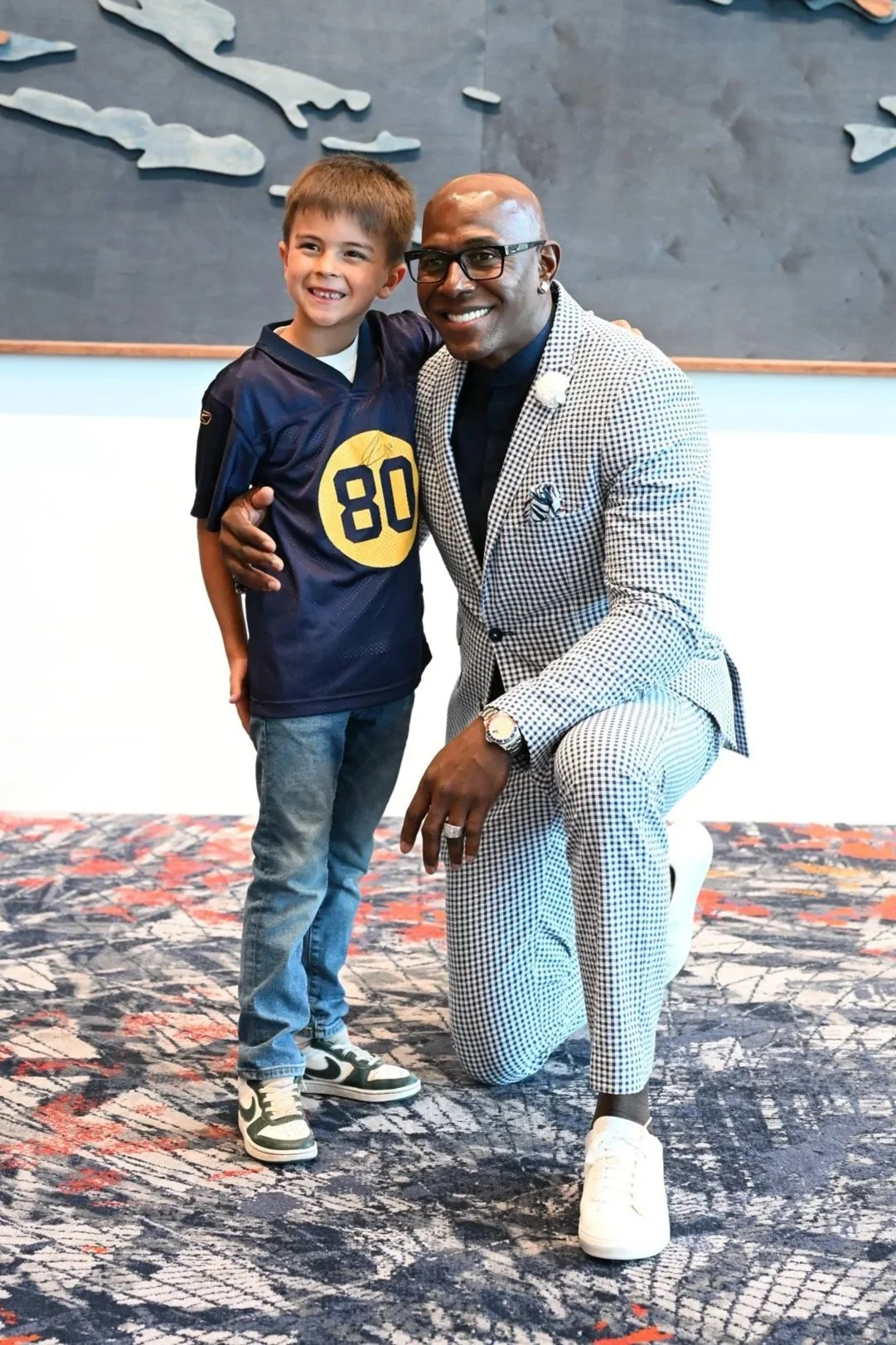 A young boy and a man are posing together indoors, both smiling. The boy is wearing a dark sports jersey with the number 80 on it, and the man is in a checkered suit with glasses, kneeling and resting one hand on his knee.