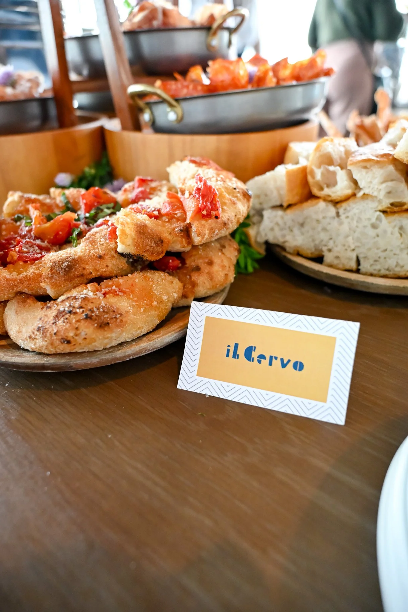 A plate of bread and a pizza with tomato sauce and herbs, placed on a wooden table with a card labeled 'il Cervo'.