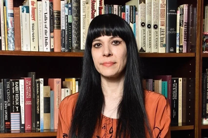 A woman with long black hair and bangs, wearing an orange top, standing in front of a bookshelf filled with various books.