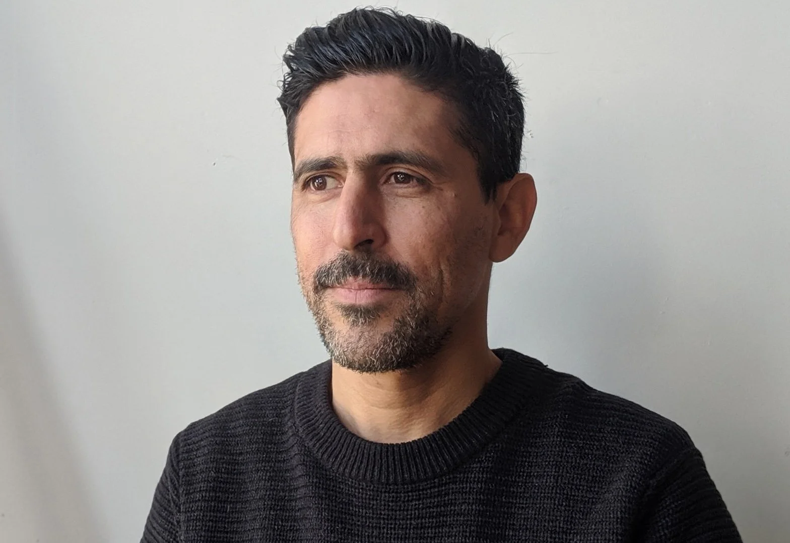 A man with dark hair, a beard, and wearing a black sweater looks to the side against a plain white background.