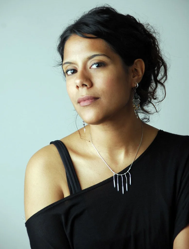 Portrait of a young woman with dark hair wearing earrings and a necklace, dressed in a black off-shoulder top, against a plain background.