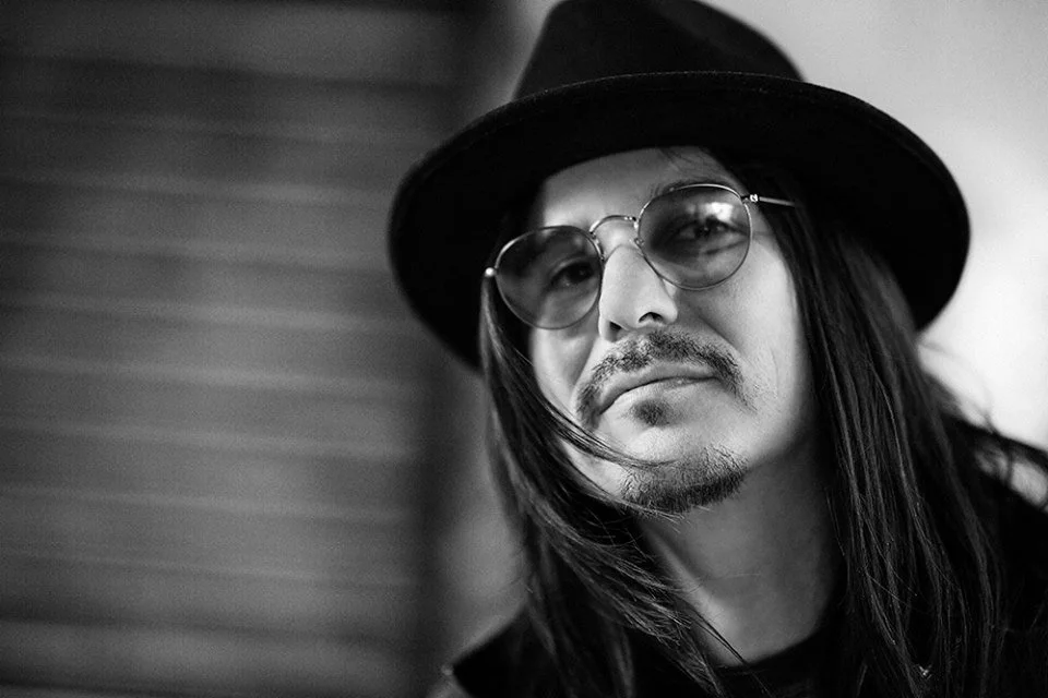 Black and white portrait of a man with long hair, wearing round glasses and a hat, looking at the camera.