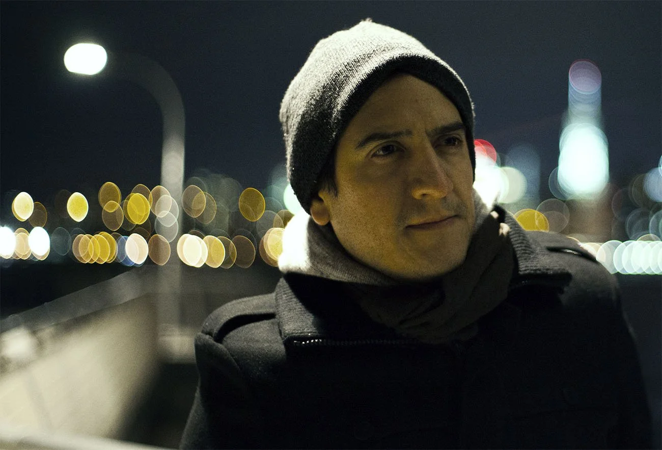 A man wearing a beanie and dark coat at night, with city lights in the background.