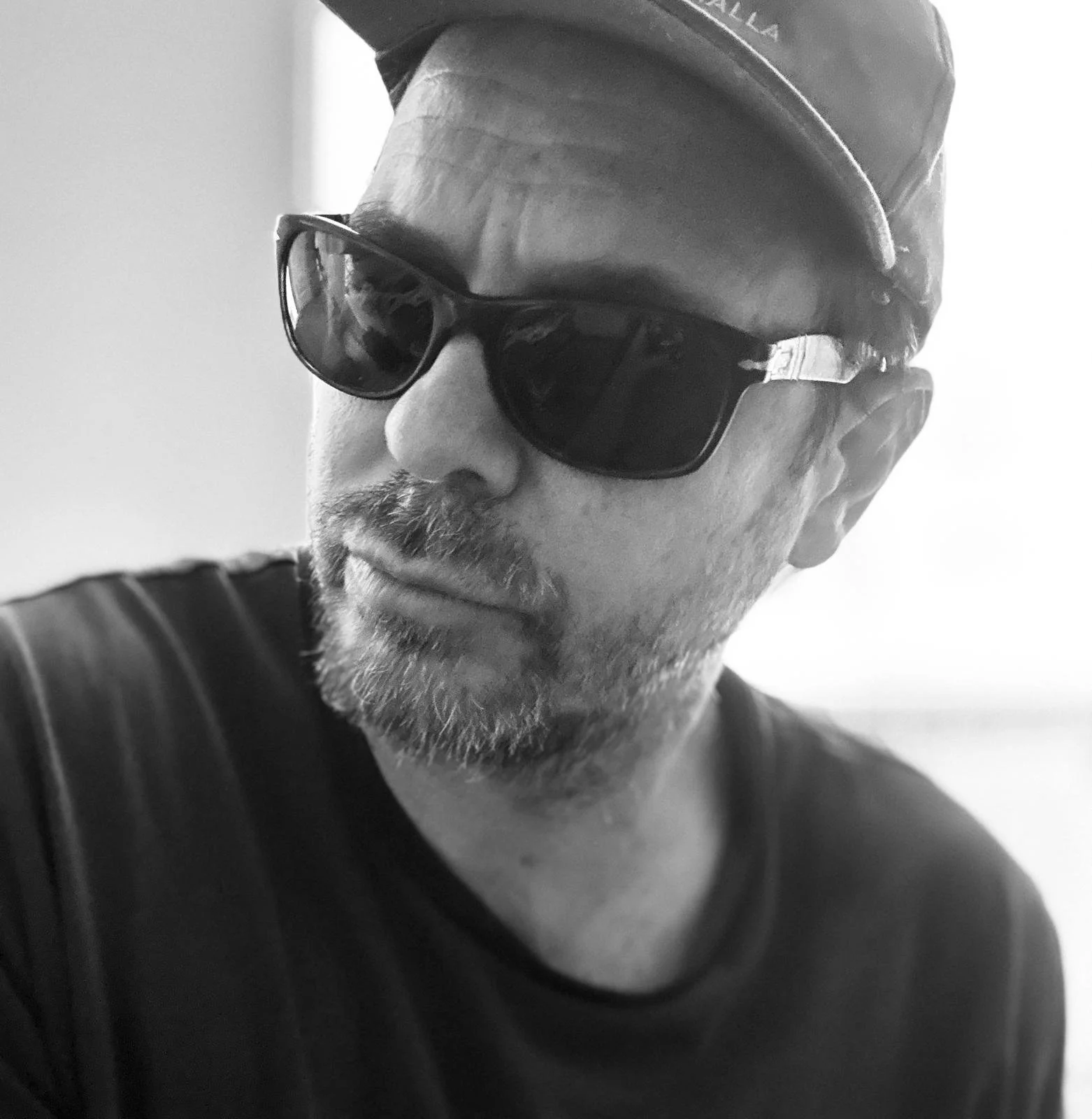 A black and white photo of a man wearing a cap and sunglasses, with a beard and mustache, looking slightly to the side.