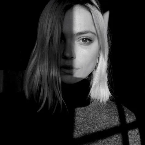 Black and white close-up photo of a young woman with shoulder-length hair, half her face lit and the other half in shadow, wearing a dark sweater.