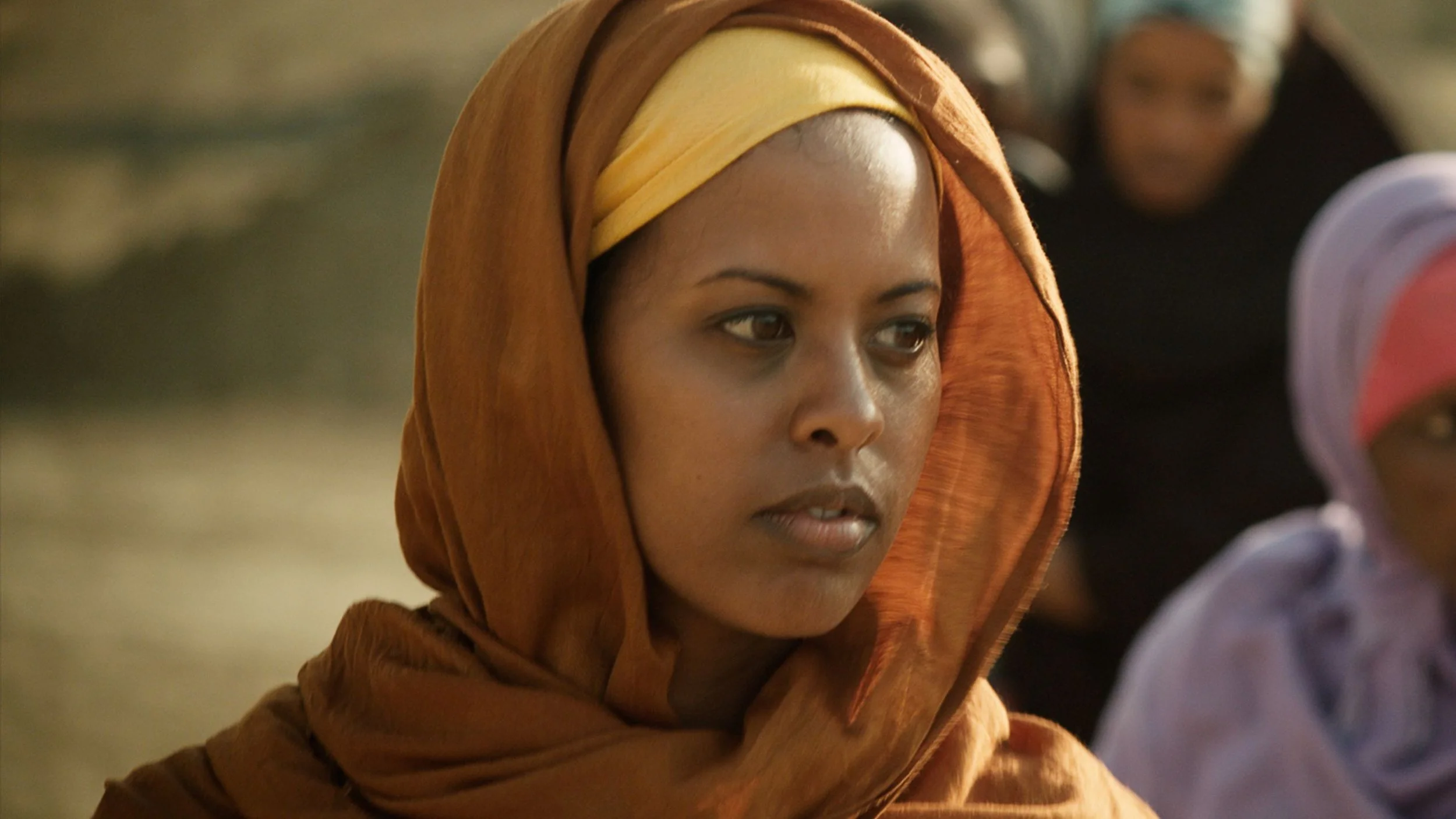 A woman wearing a brown headscarf and yellow head covering sitting outdoors with other women in the background.