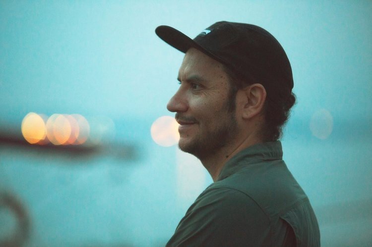 A man with a beard and a baseball cap standing outdoors at dusk, with blurred city lights in the background.