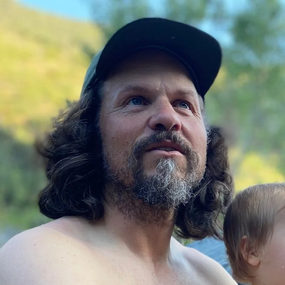 A shirtless man with long dark hair, a beard with gray, wearing a black cap, outdoors with blurred trees in the background.