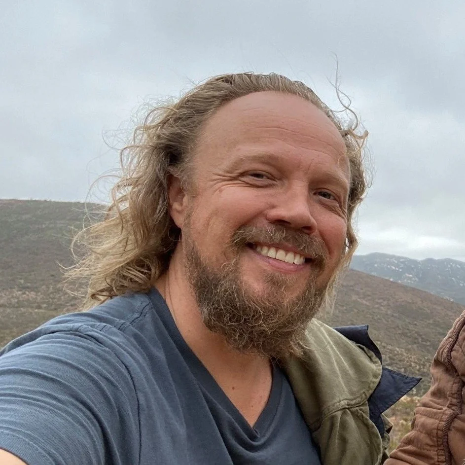 Smiling man with long hair and beard outdoors, mountain landscape background.