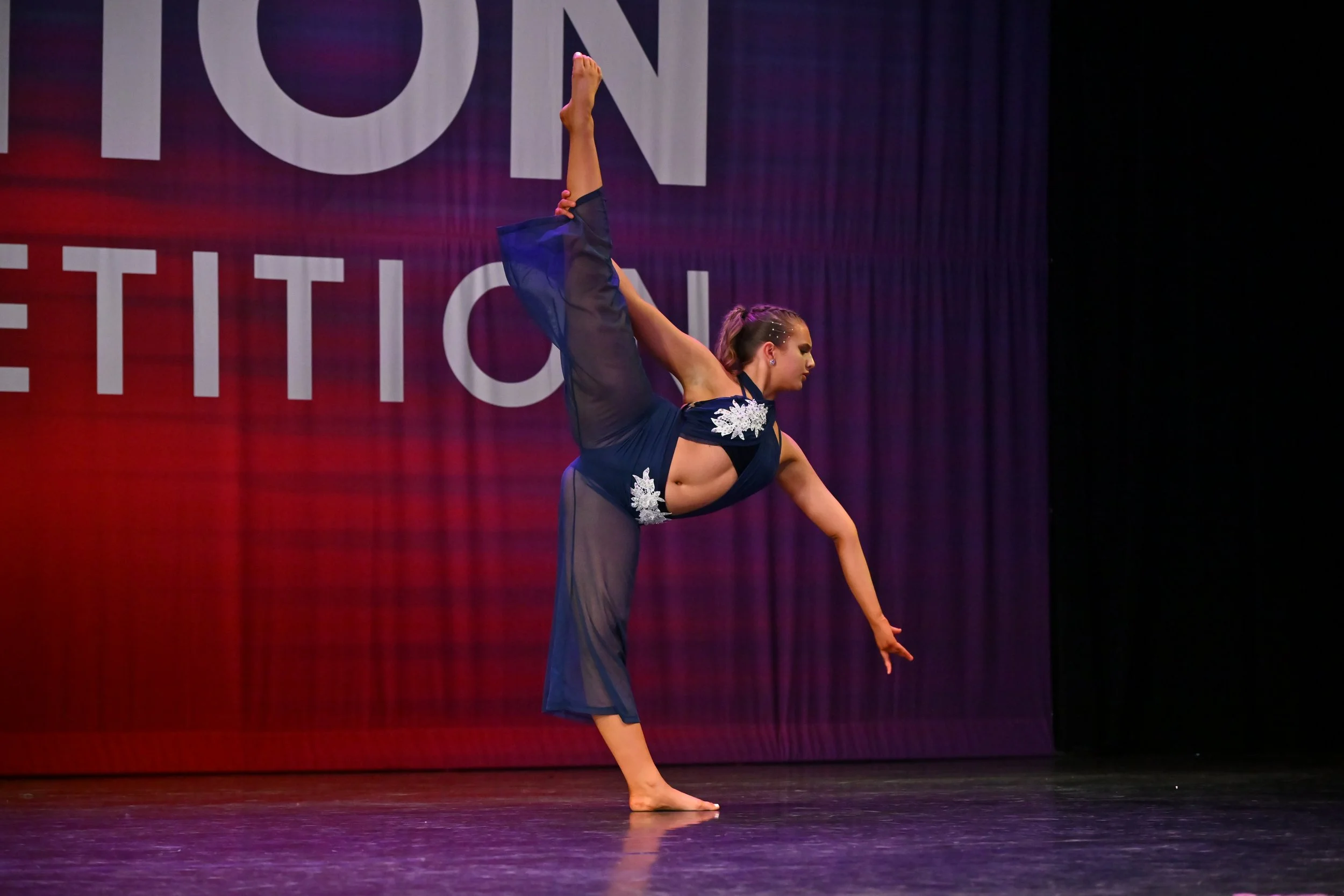 A female dancer performing a split on stage while balancing on one hand with her leg extended upward during a dance competition.