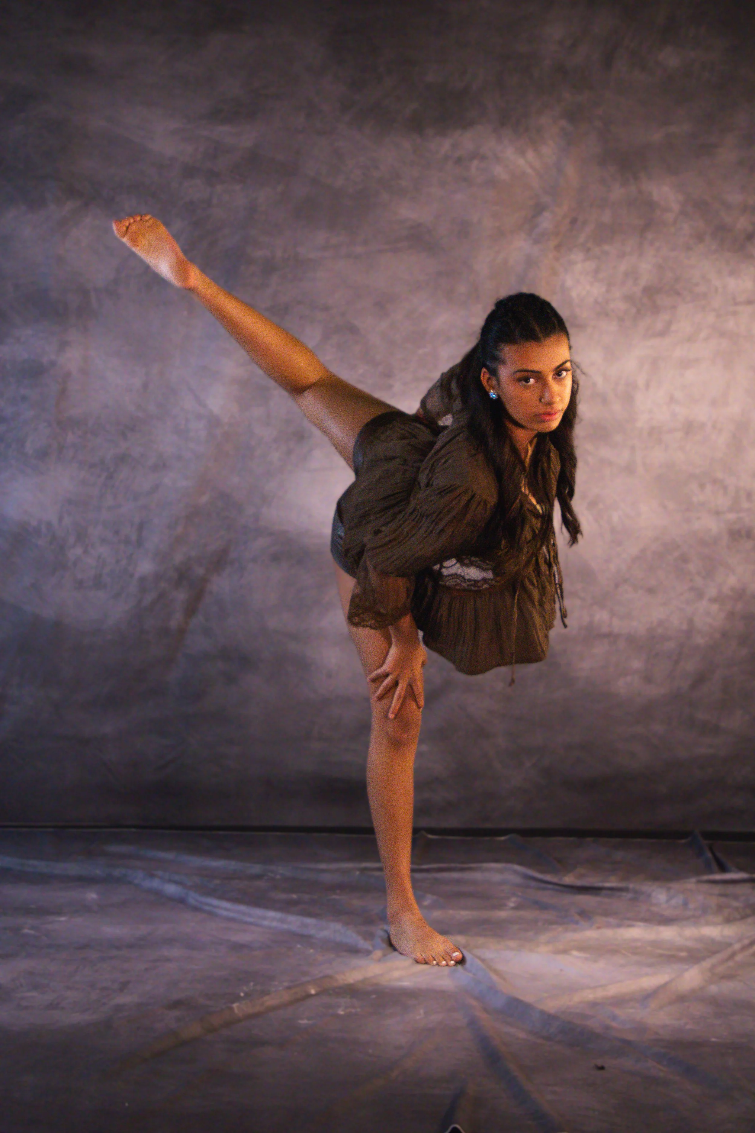 A woman in a dancer pose standing on one leg with the other leg extended backwards, one hand on her bent knee, wearing a brown dress and blue earrings, set against a dark, textured background.