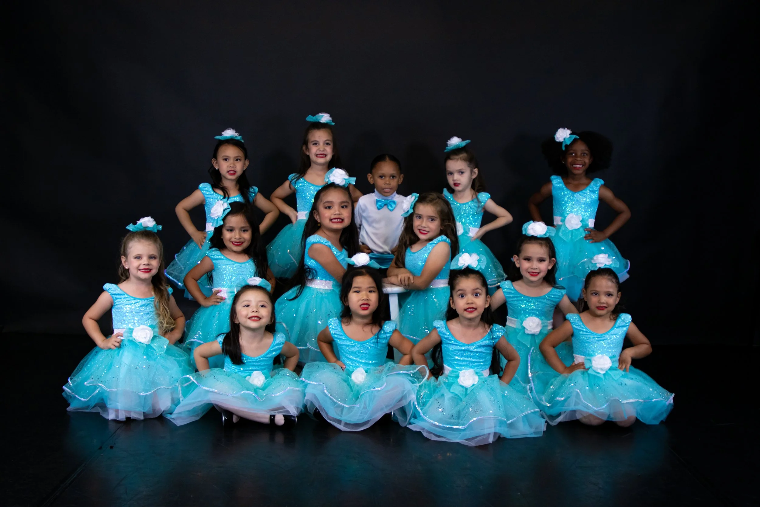 A group of young girls in matching turquoise dance costumes with white accents posing together on a black background.