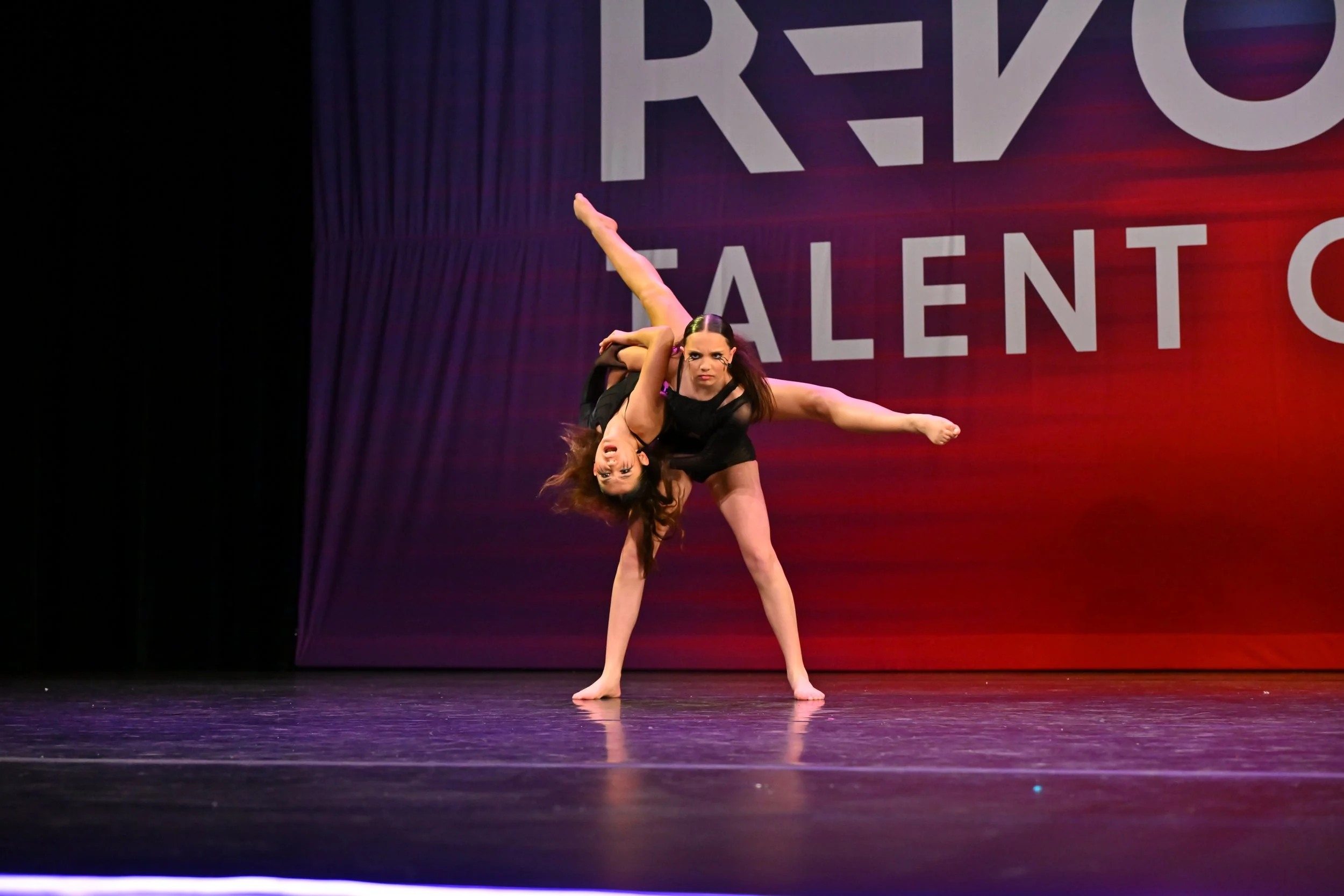 Two dancers performing a contemporary dance routine on stage, one lifting the other while balancing on one leg, with a large sign in the background reading 'RARE TALENT'.