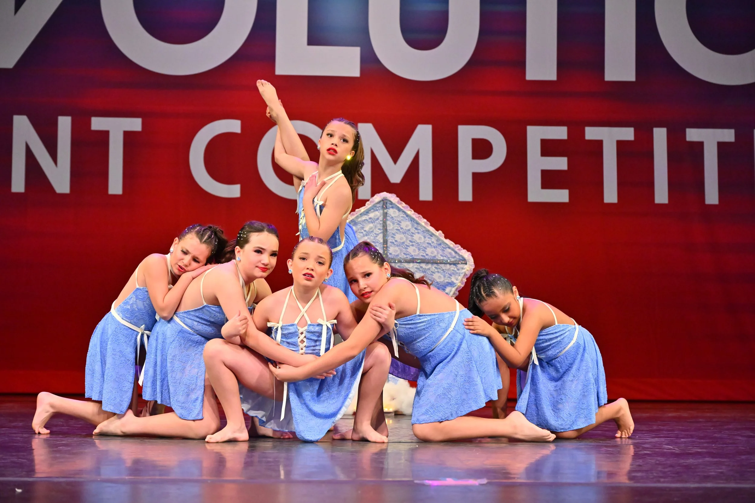 A group of young girls dressed in blue costumes performing a dance on stage with a red backdrop and large text.