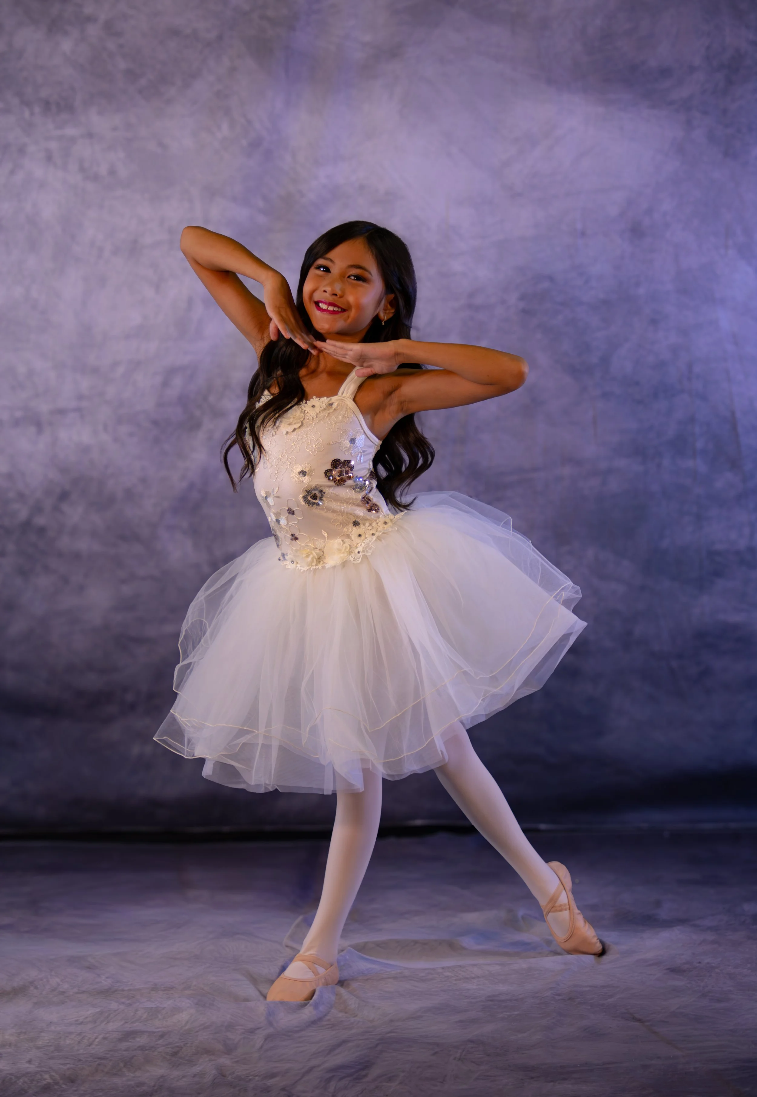A young girl in a white tutu dress with floral embroidery and beige ballet shoes poses as a ballet dancer against a textured purple background.