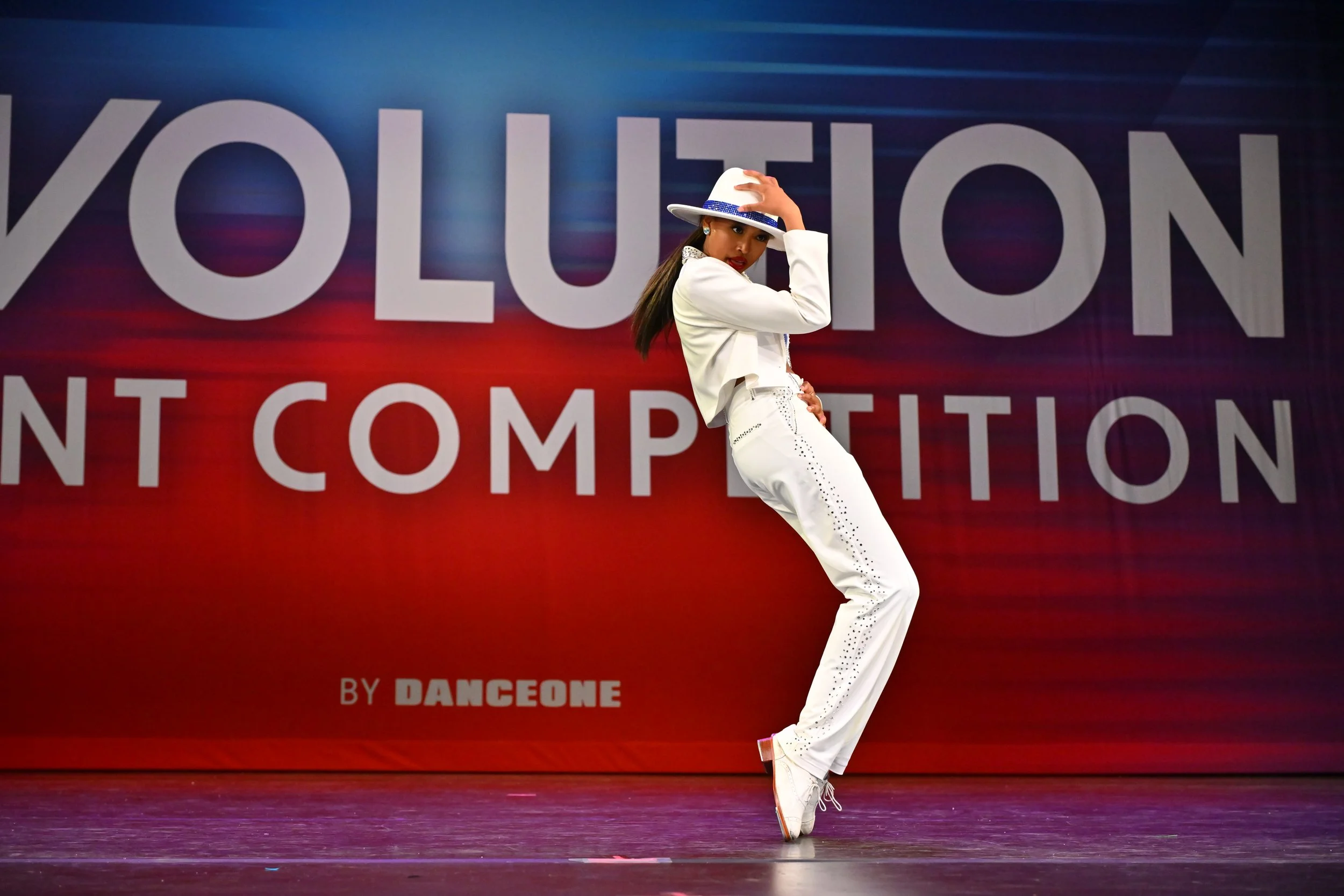 A female dancer in white pants and a cropped jacket posing with one hand on her hat against a backdrop with large text 'VOLUTION' and smaller text 'BY DANCEONE'.