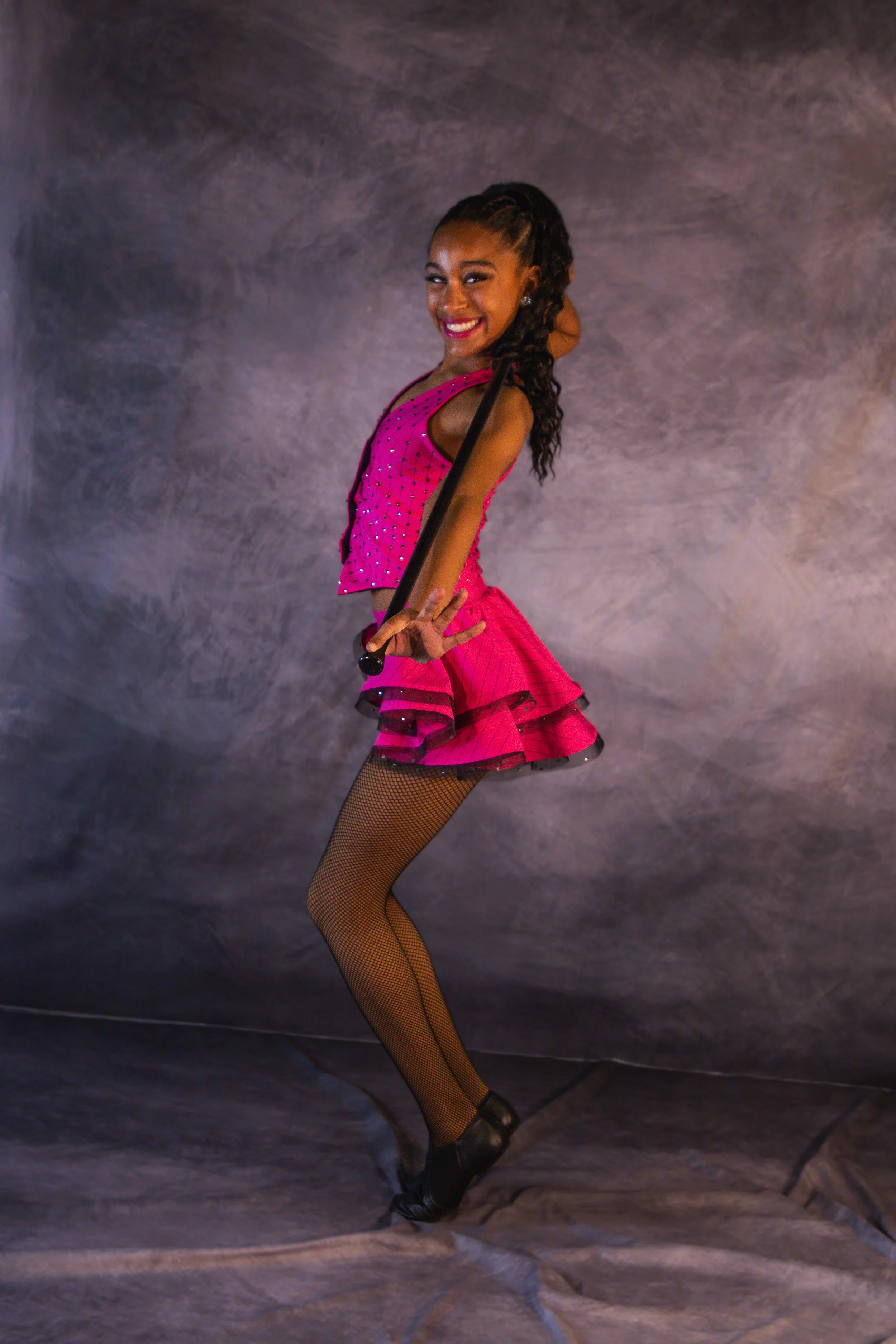 A woman in a pink dance costume with sparkles, fishnet stockings, and black dance shoes, posing in front of a textured gray background.