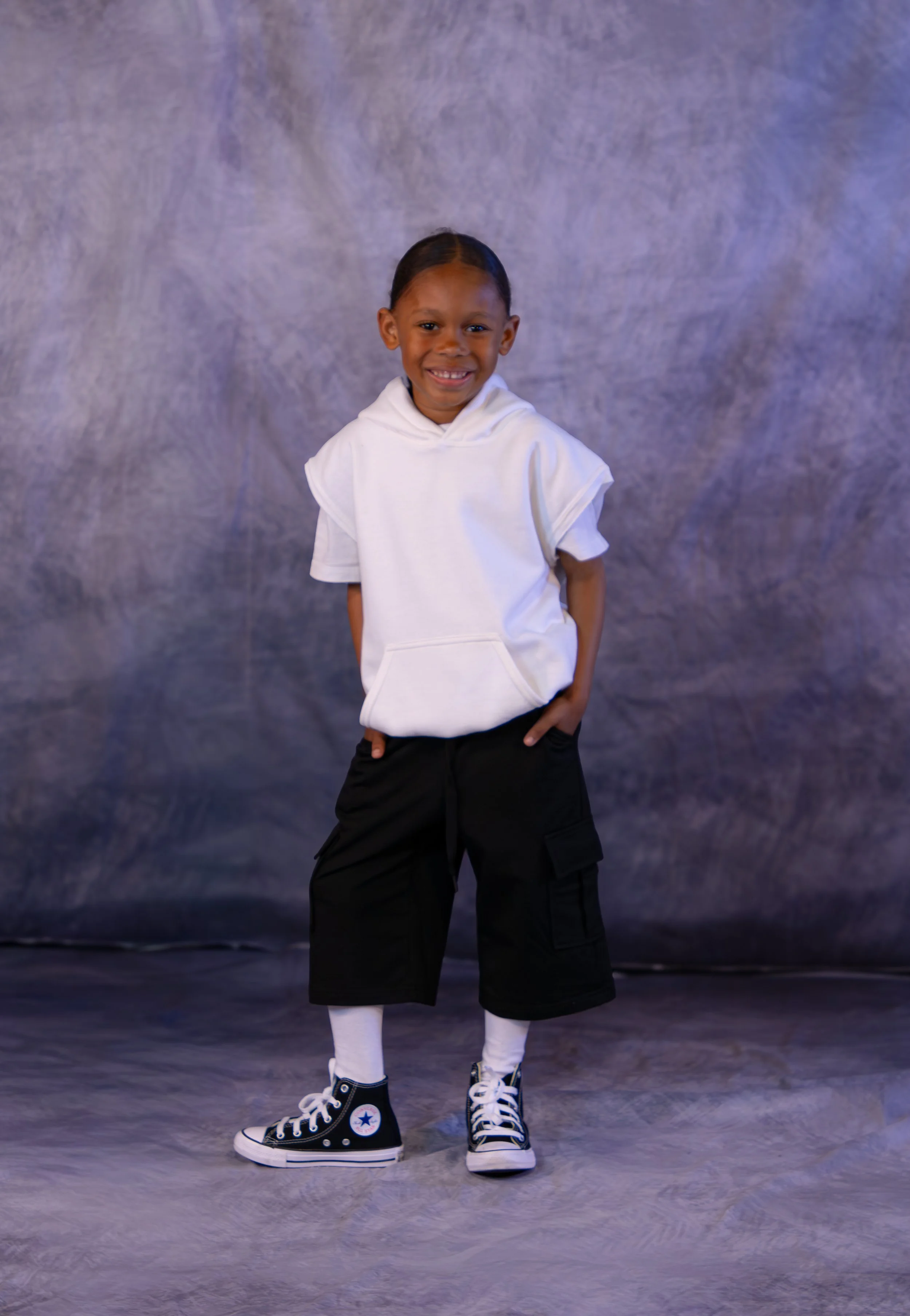 A young boy smiling, standing with hands in pockets, wearing a white hoodie, black cargo shorts, white socks, and black Converse sneakers, against a mottled gray backdrop.
