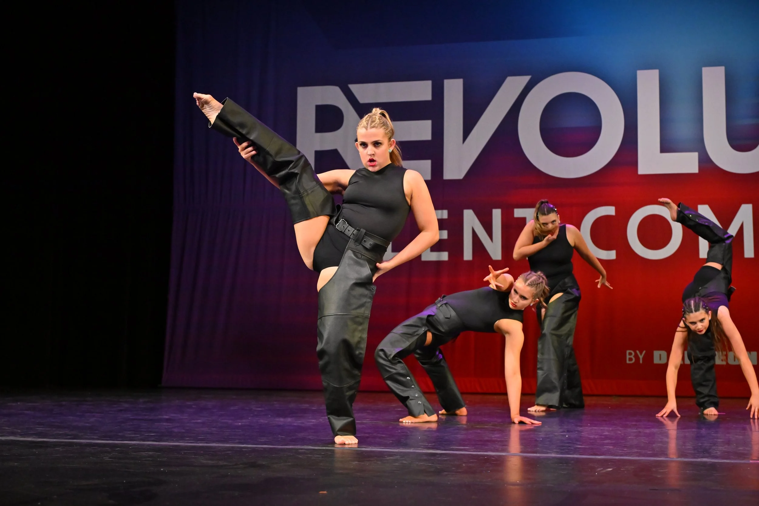 A group of female dancers performing a dance routine on stage, with one dancer in the foreground executing a high kick during their routine, against a backdrop that reads 'REYOU'.