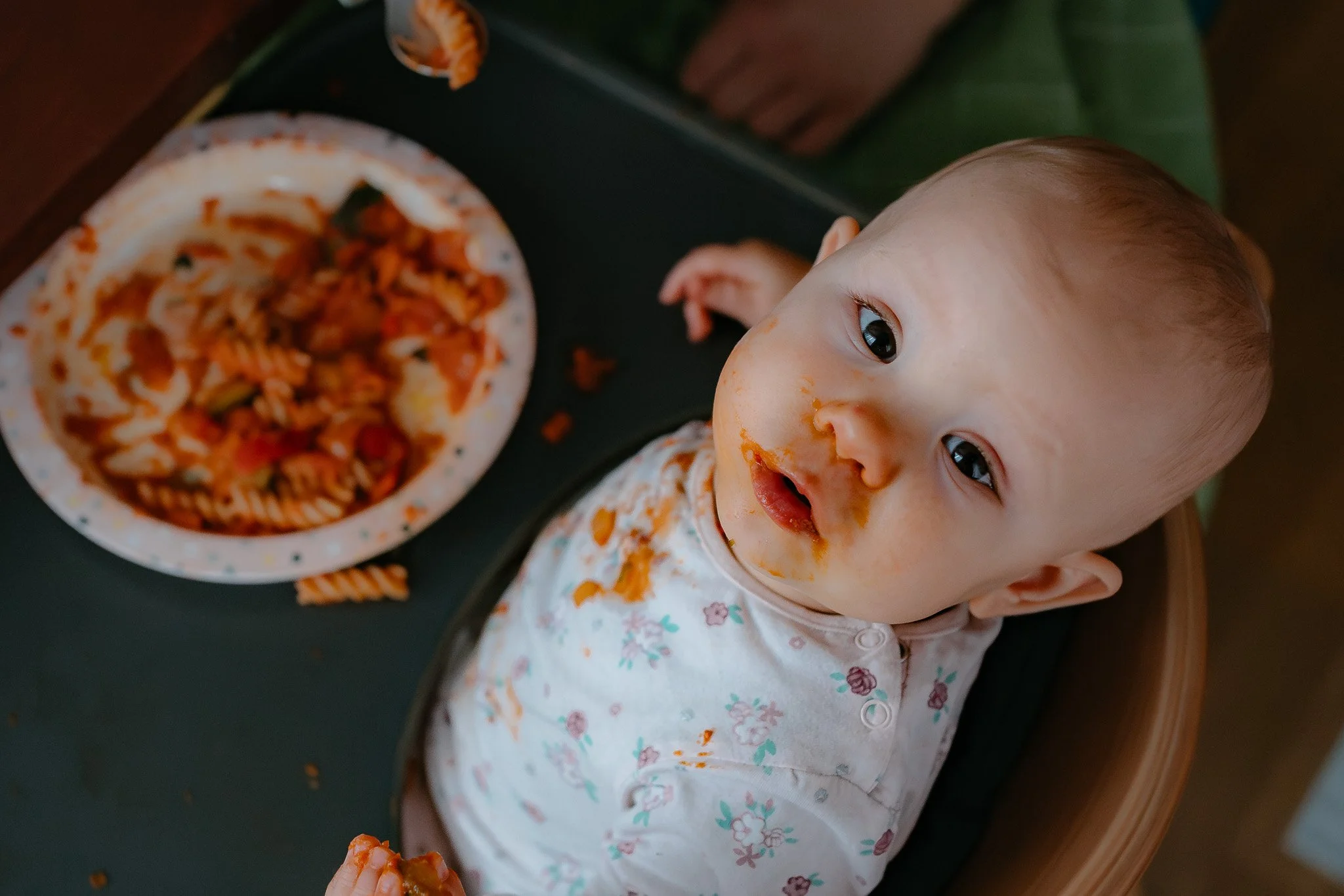 Ein junges Baby sitzt in einem Hochstuhl mit restlicher Pasta im Mund und auf dem T-Shirt, neben einem Teller mit ungegartem Pasta-Gericht.