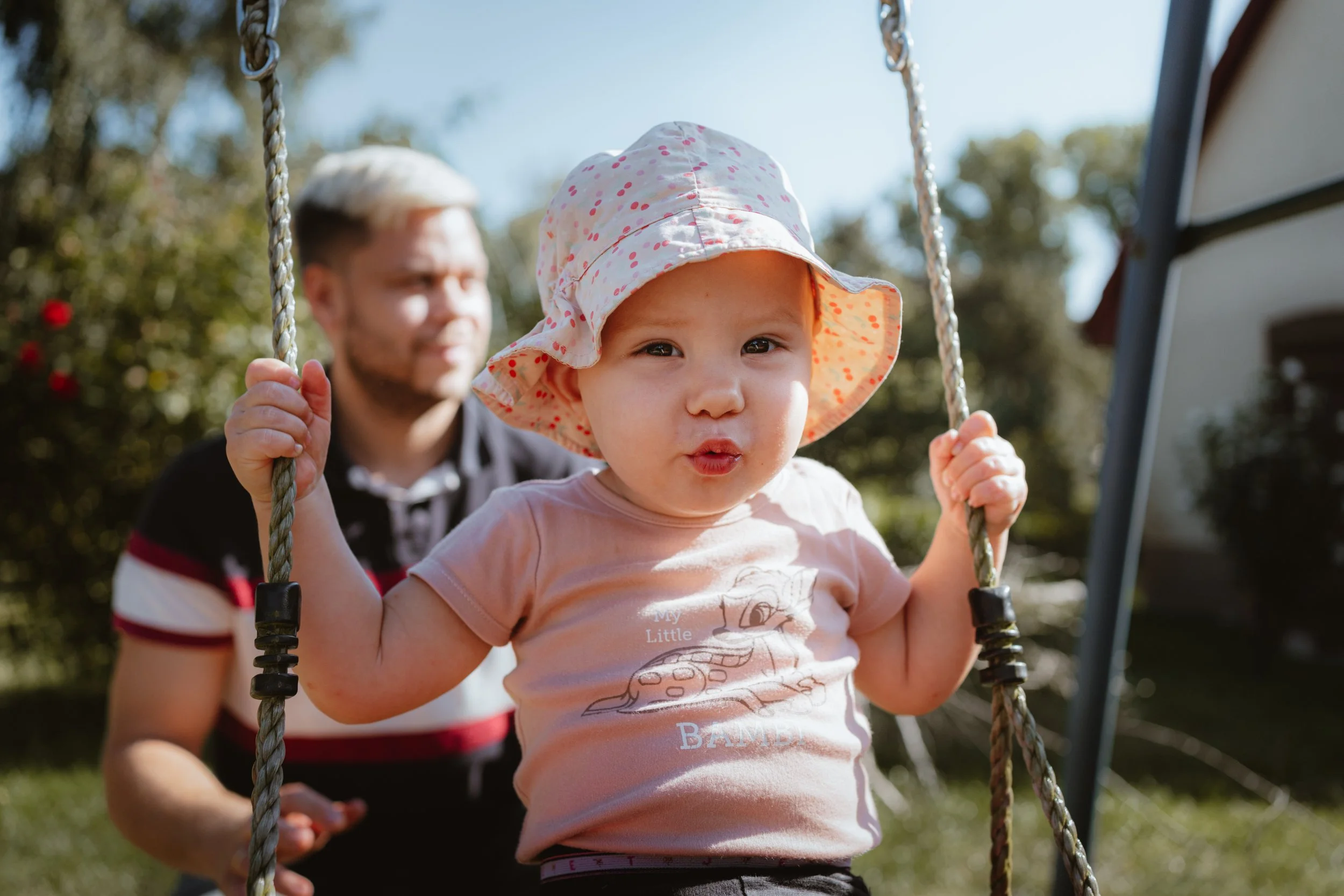 Kleines Mädchen trägt einen Sonnenhut, sitzt auf einer Schaukel im Garten, während ein Mann (wahrscheinlich ihr Vater) im Hintergrund steht.