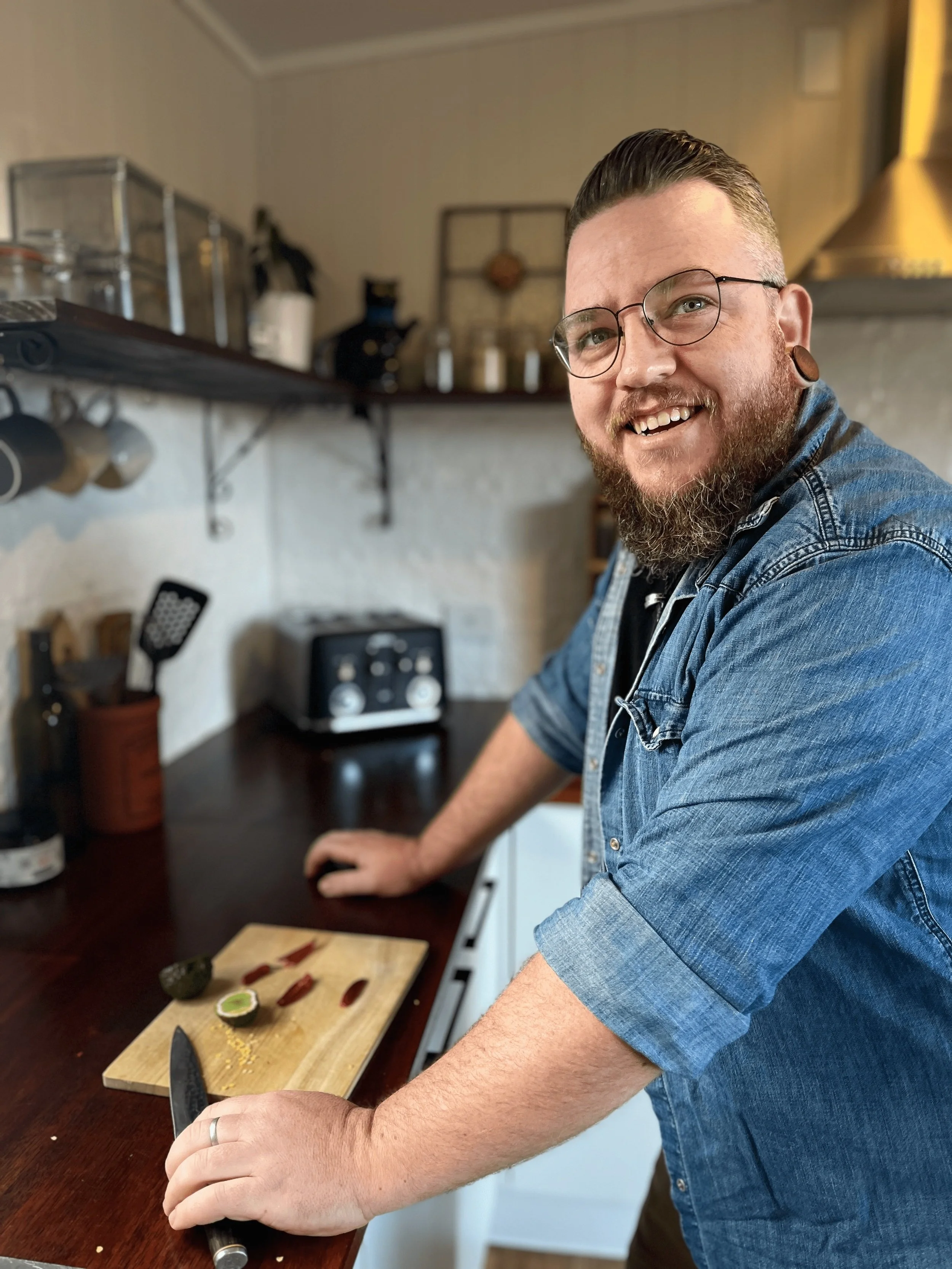 A smiling man with glasses and a beard preparing food in a kitchen, standing at a wooden counter with a cutting board and a knife, with a lime, hot peppers, and avocado nearby.