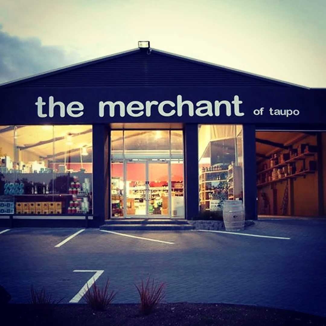 Front view of a store called 'the merchant of Taupo' with large glass windows displaying products inside, in a parking lot with marked parking spaces and small plants in the foreground.