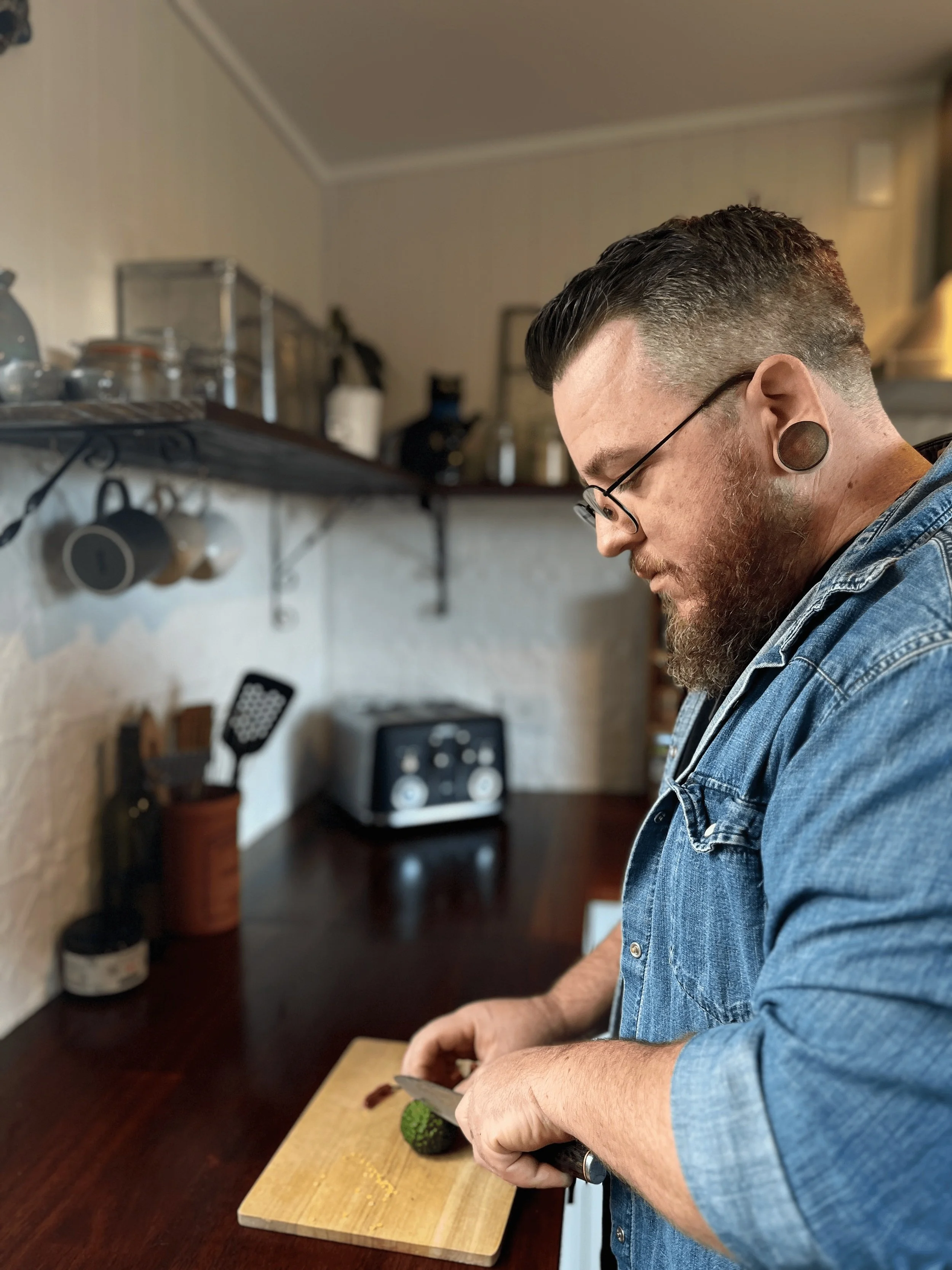 A man with glasses and gauges in his ears chopping an avocado on a wooden cutting board in a kitchen.