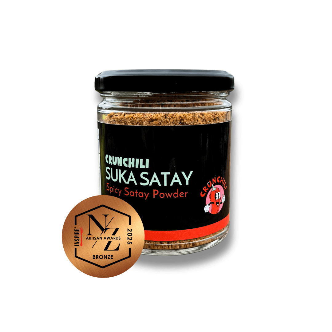 A glass jar of Crunchilli Suka Satay spicy satay powder with a black lid, black label, red accents, and a cartoon chili pepper logo. An award badge indicating a bronze prize in the 2025 Inspire+ Artisan Awards is attached.