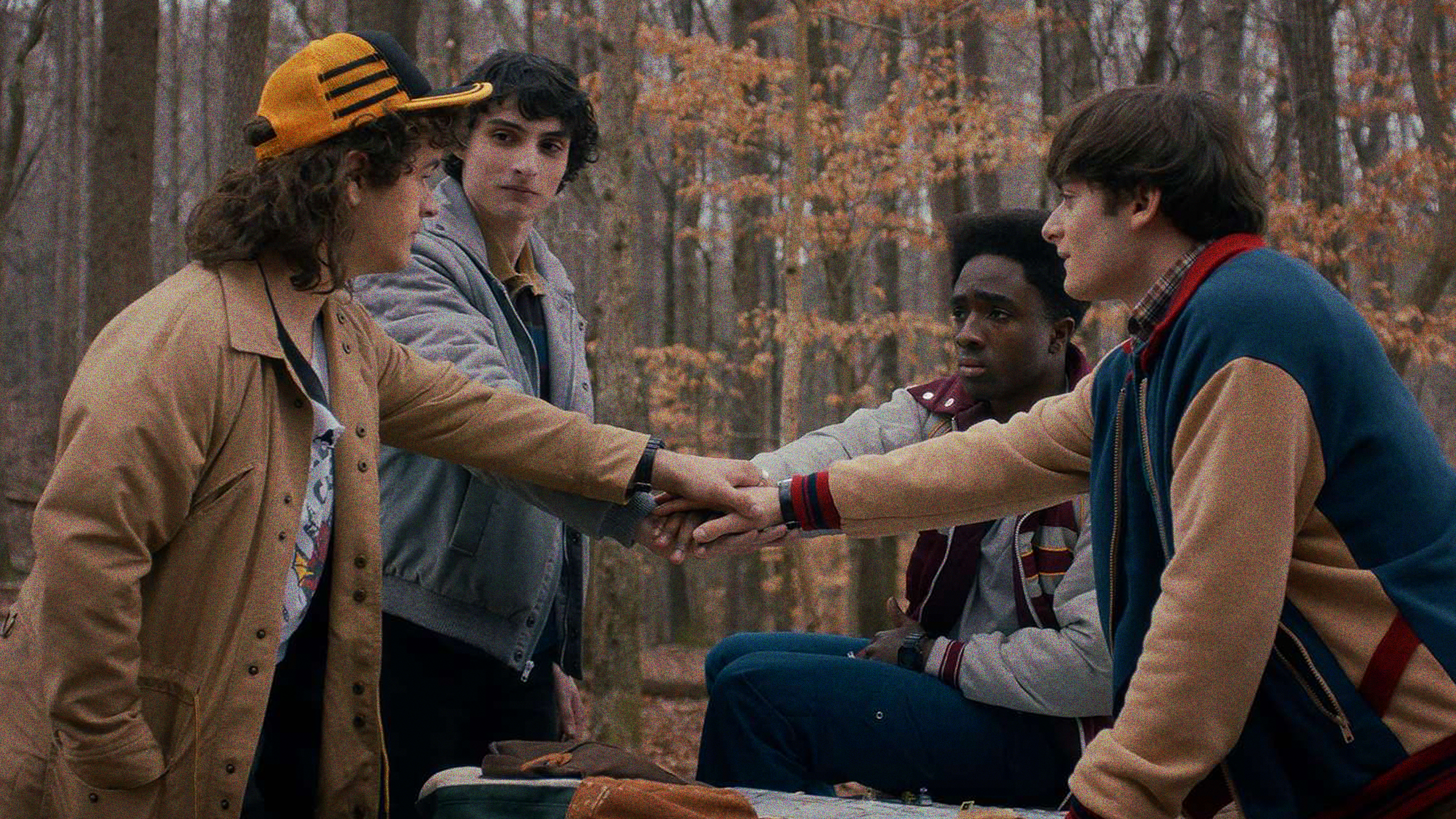 The characters Dustin, Will, Lucas and Mike of Stranger things holding hands in a group in the middle of a forest in the fall