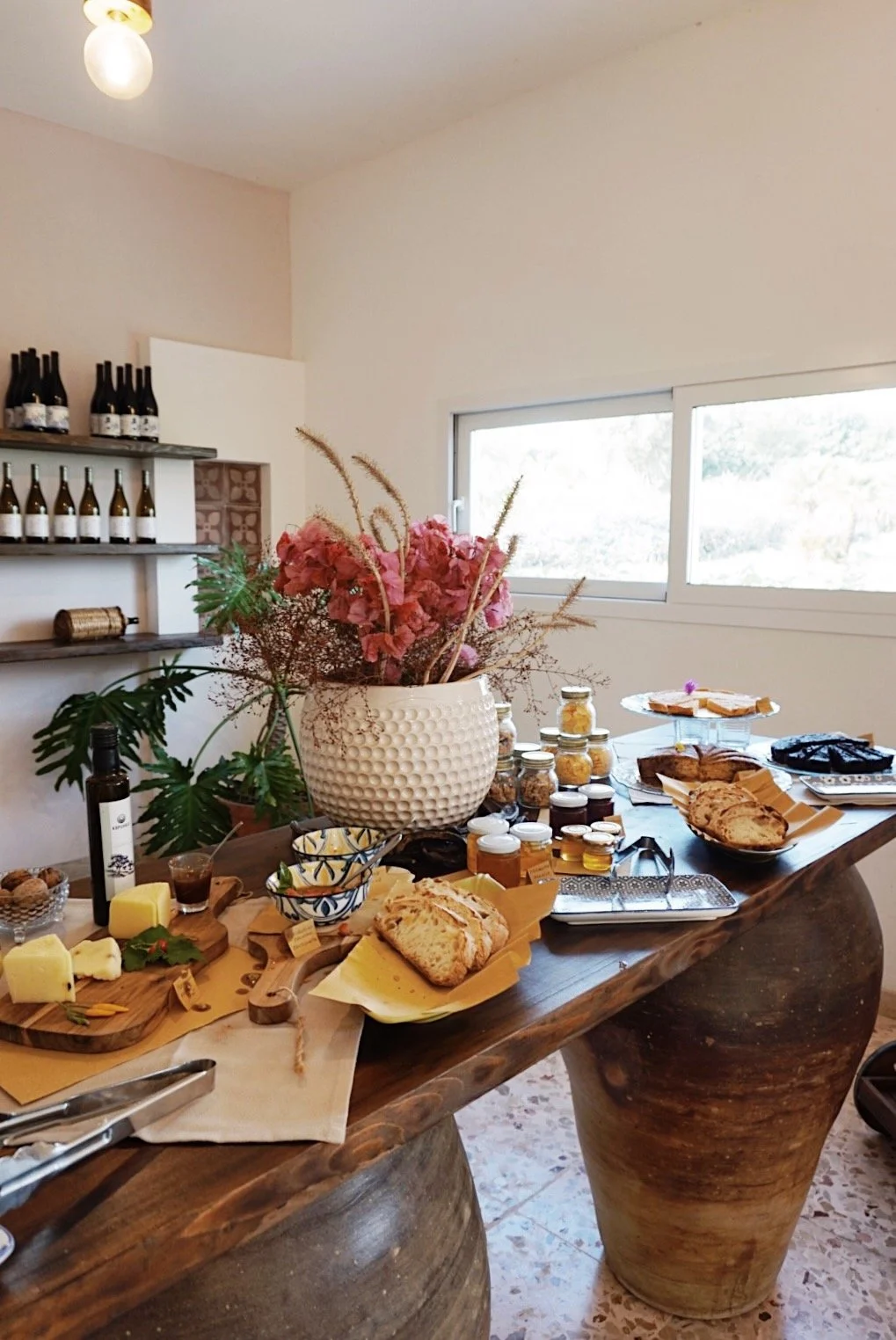 Buffet table with assorted breads, cheeses, and jars of honey, decorative flowers in a white vase, shelf with wine bottles, and a window view.