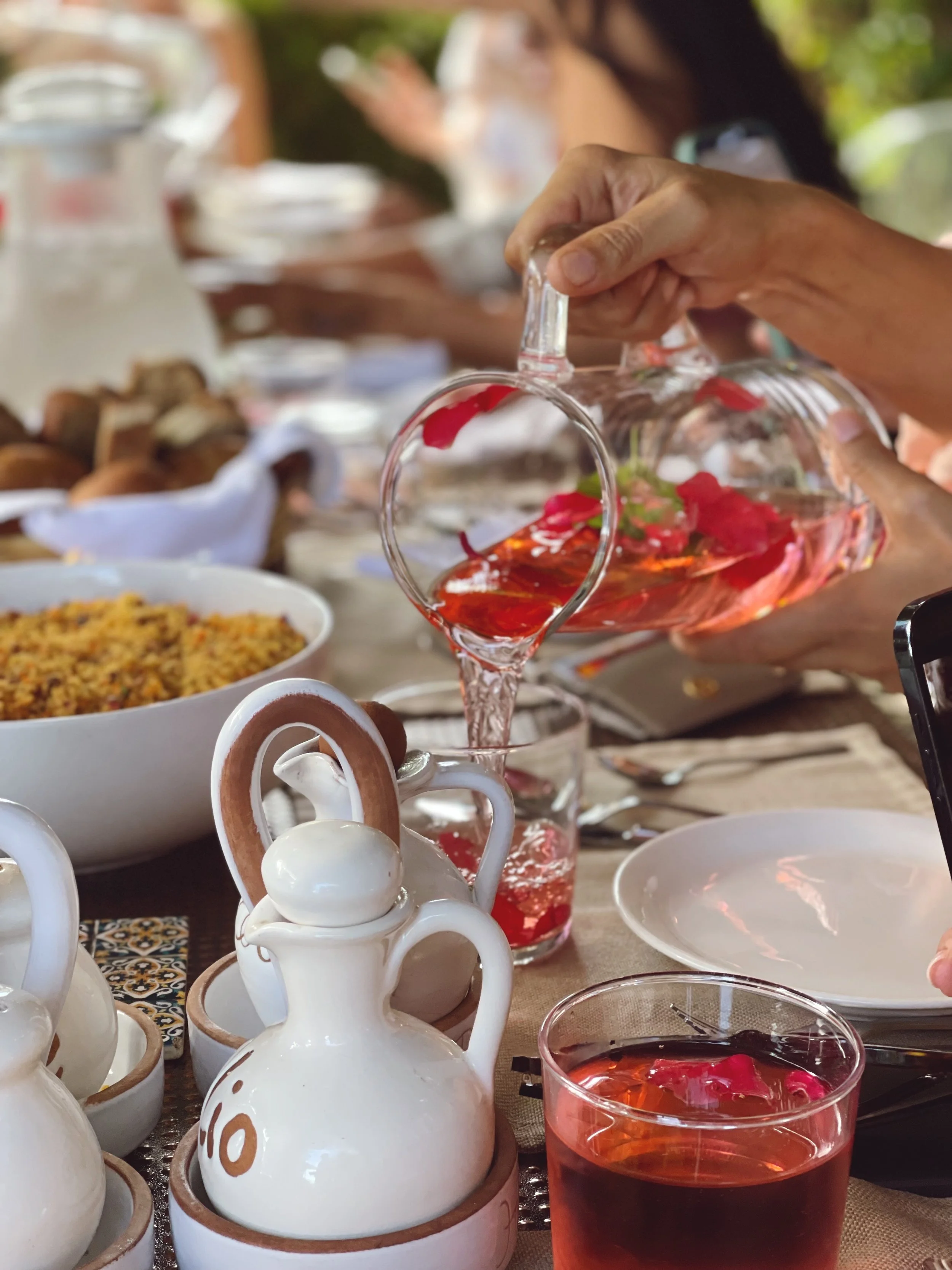A person pouring red liquid from a glass pitcher into a glass on a dining table. The table has white ceramic jugs, a bowl of food, and a glass of red drink with flower petals. Plates and cutlery are also visible.