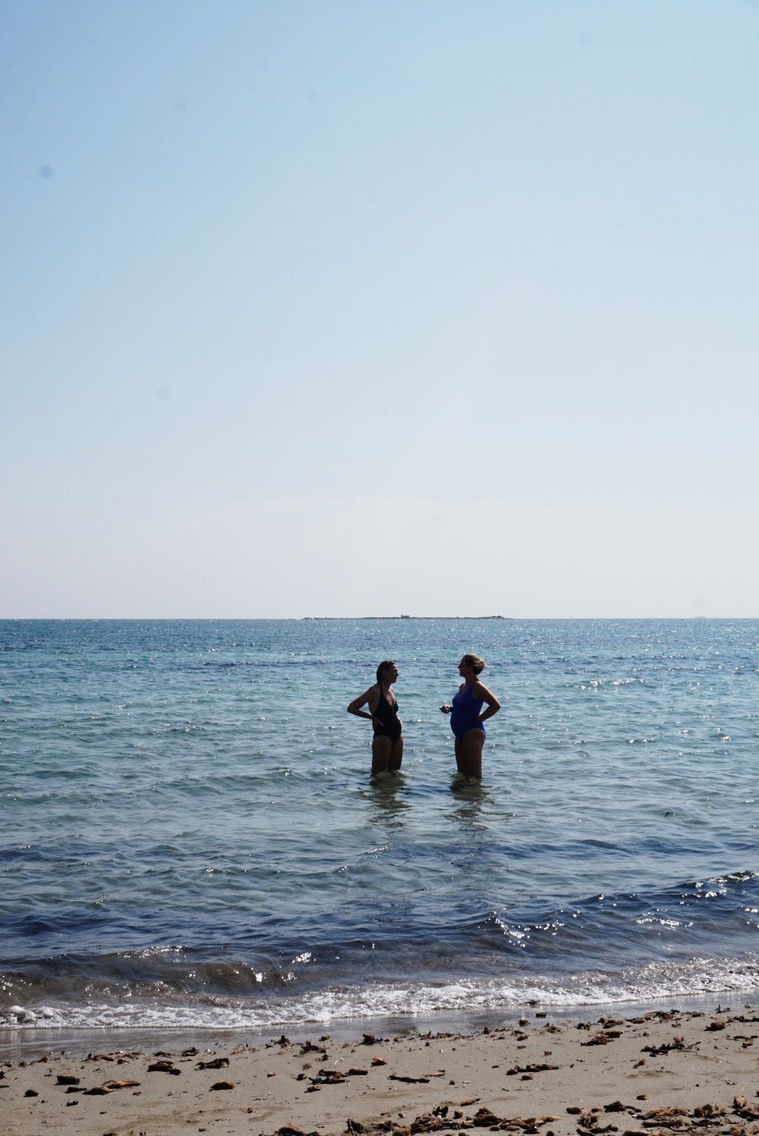 Two people standing in shallow ocean water near a sandy beach, conversing.