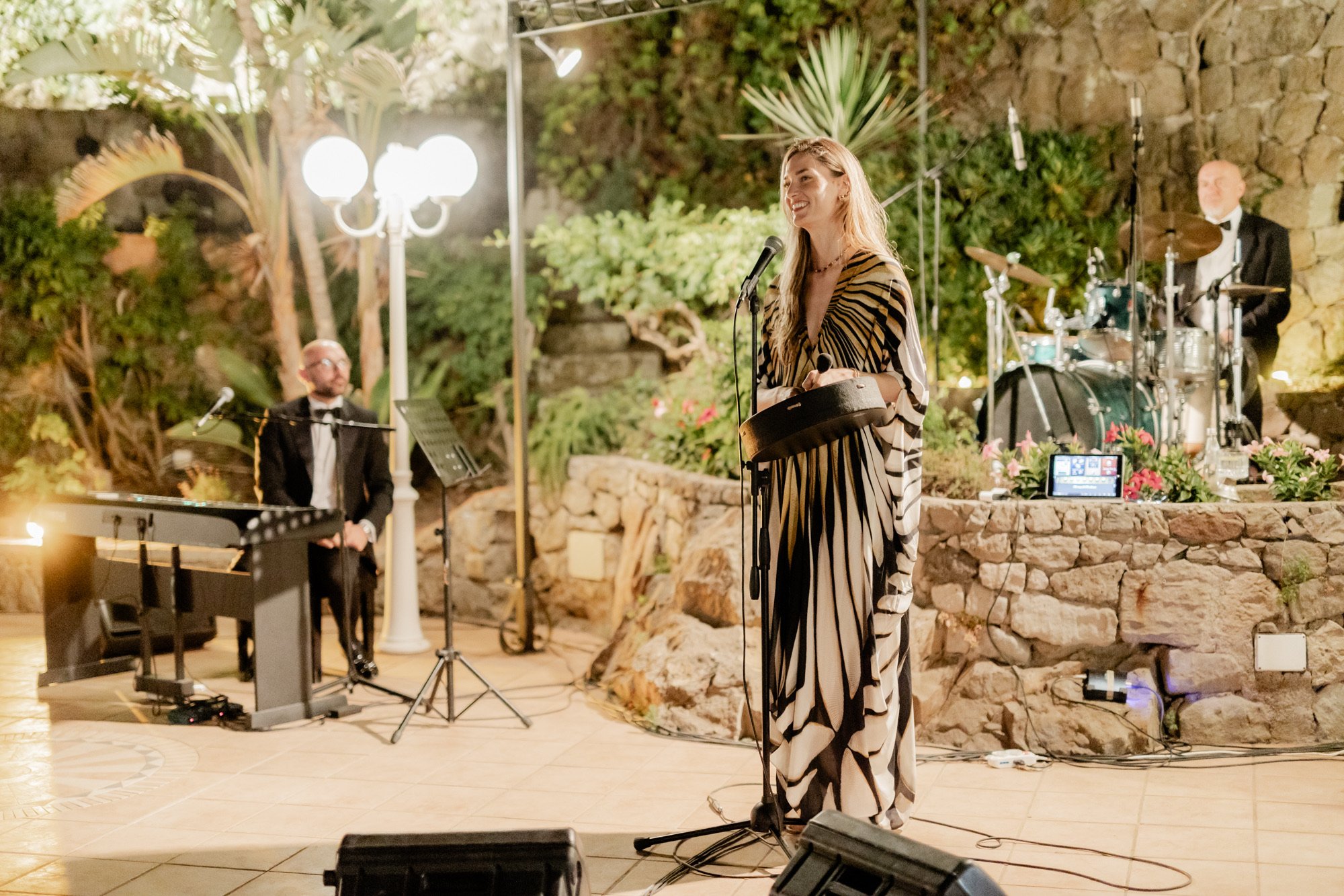 A singer performing outdoors with a musician on keyboard and a drummer in the background. The setting includes stone walls and lush greenery.