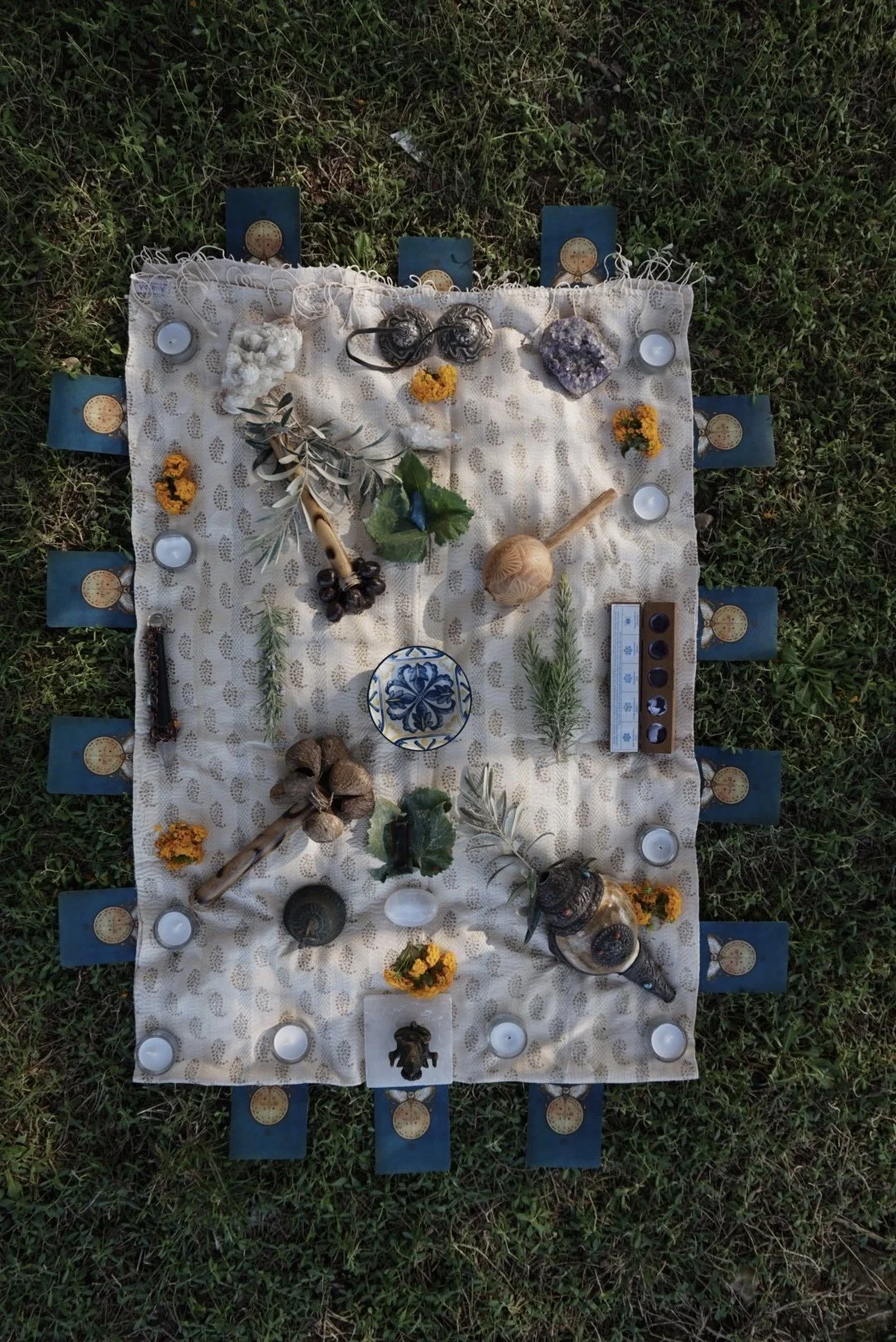 A ritual setup on a cloth laid on grass with various objects including crystals, candles, herbs, and decorative items arranged symmetrically.