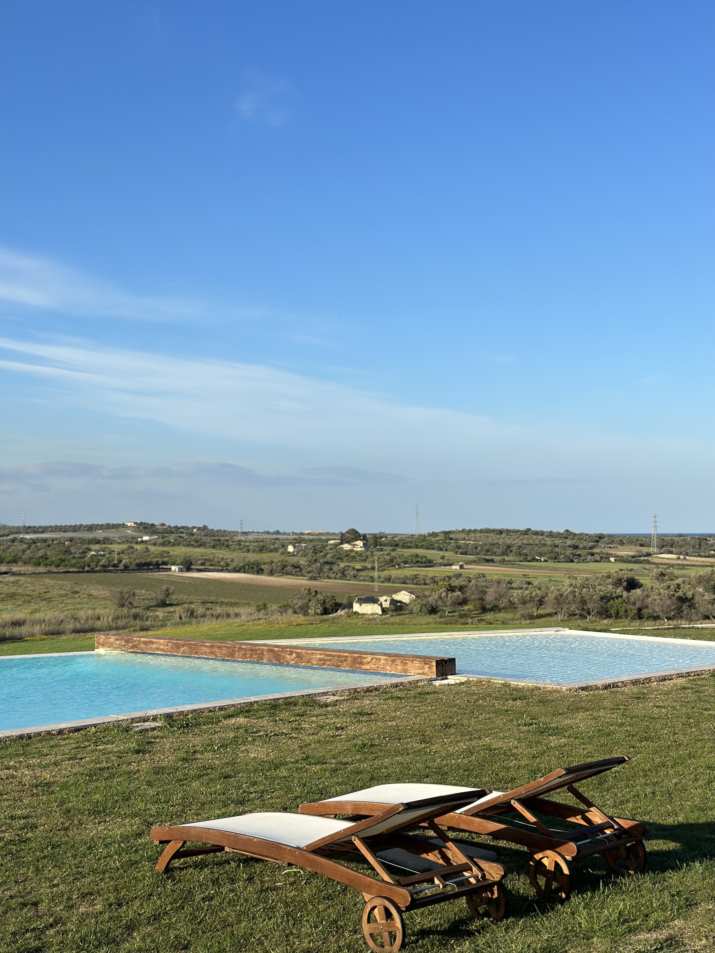 Two wooden lounge chairs on a grassy lawn overlooking an infinity pool with a scenic view of rolling hills and a clear blue sky.