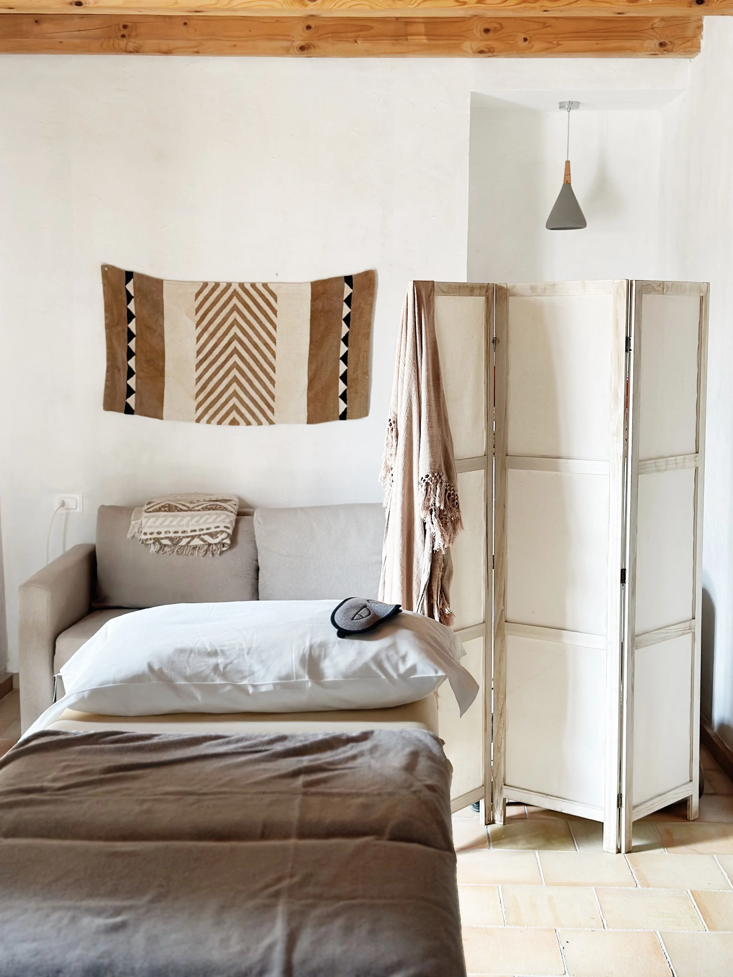 Cozy interior with a bed, beige couch, and a room divider. A stylish textile hangs on the wall, and there's a pendant light overhead. Neutral colors and simple decor create a relaxing ambiance.