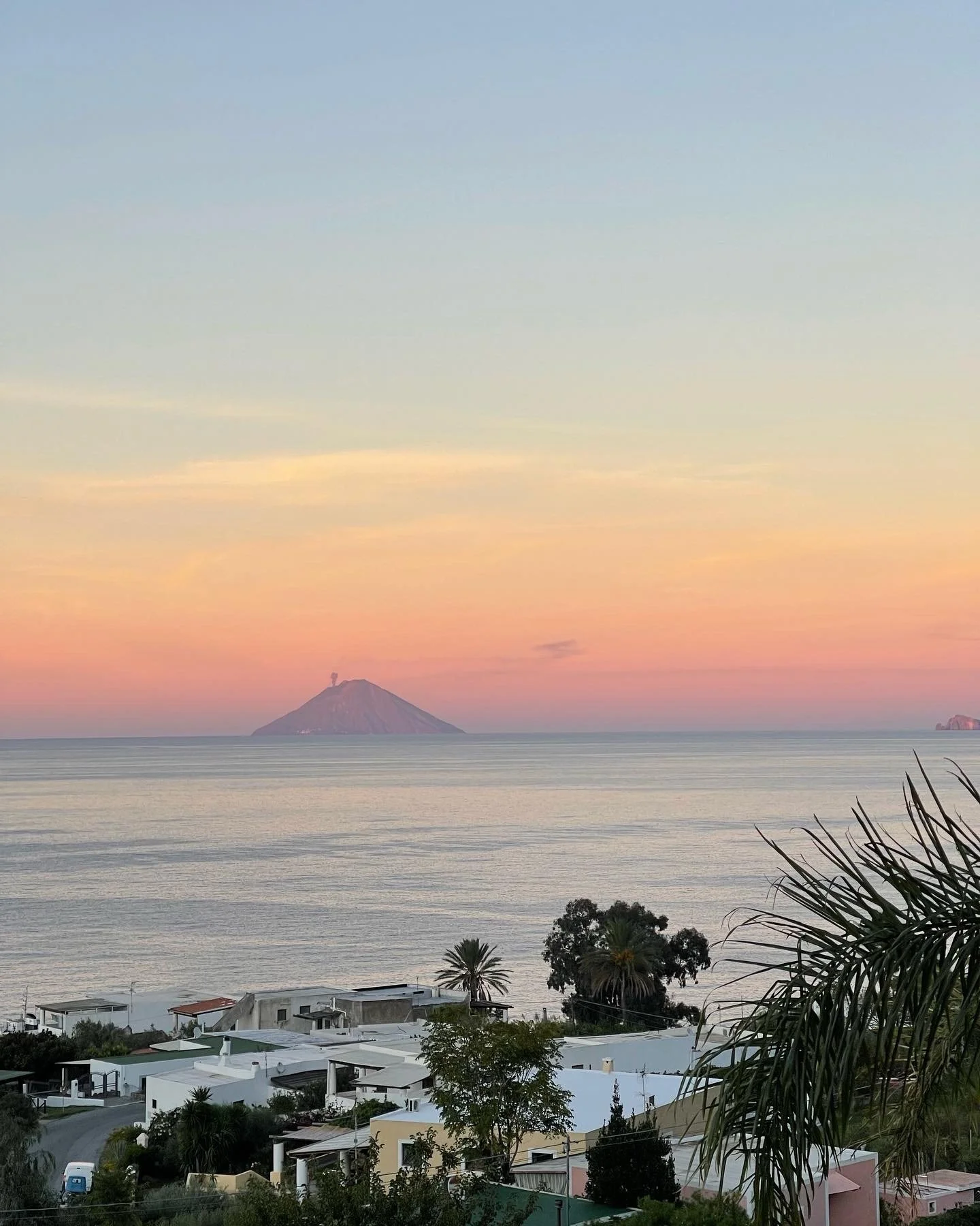 View of a distant island with a volcano, sunset sky, foreground with trees, rooftops, and a palm tree overlooking the ocean.