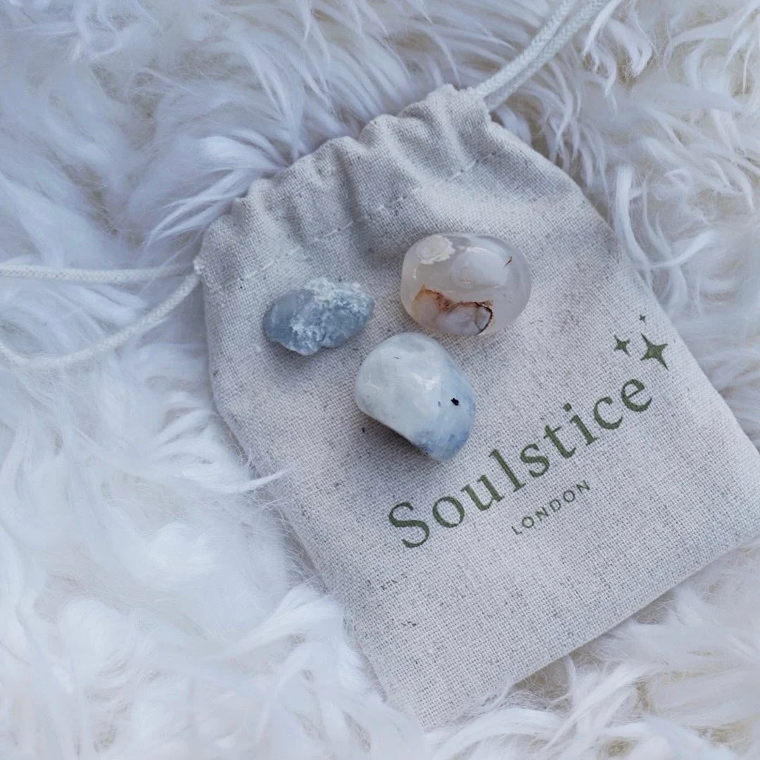Gray drawstring pouch with "Soulstice London" text, containing three polished stones, on a white fluffy background.