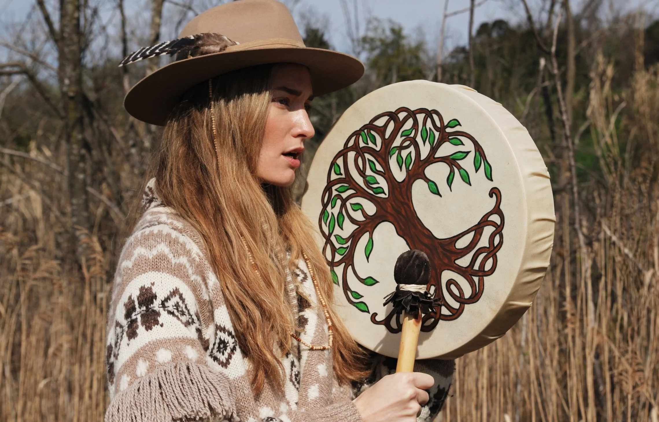 Person in hat and patterned sweater playing a hand drum with a tree design in a natural setting.