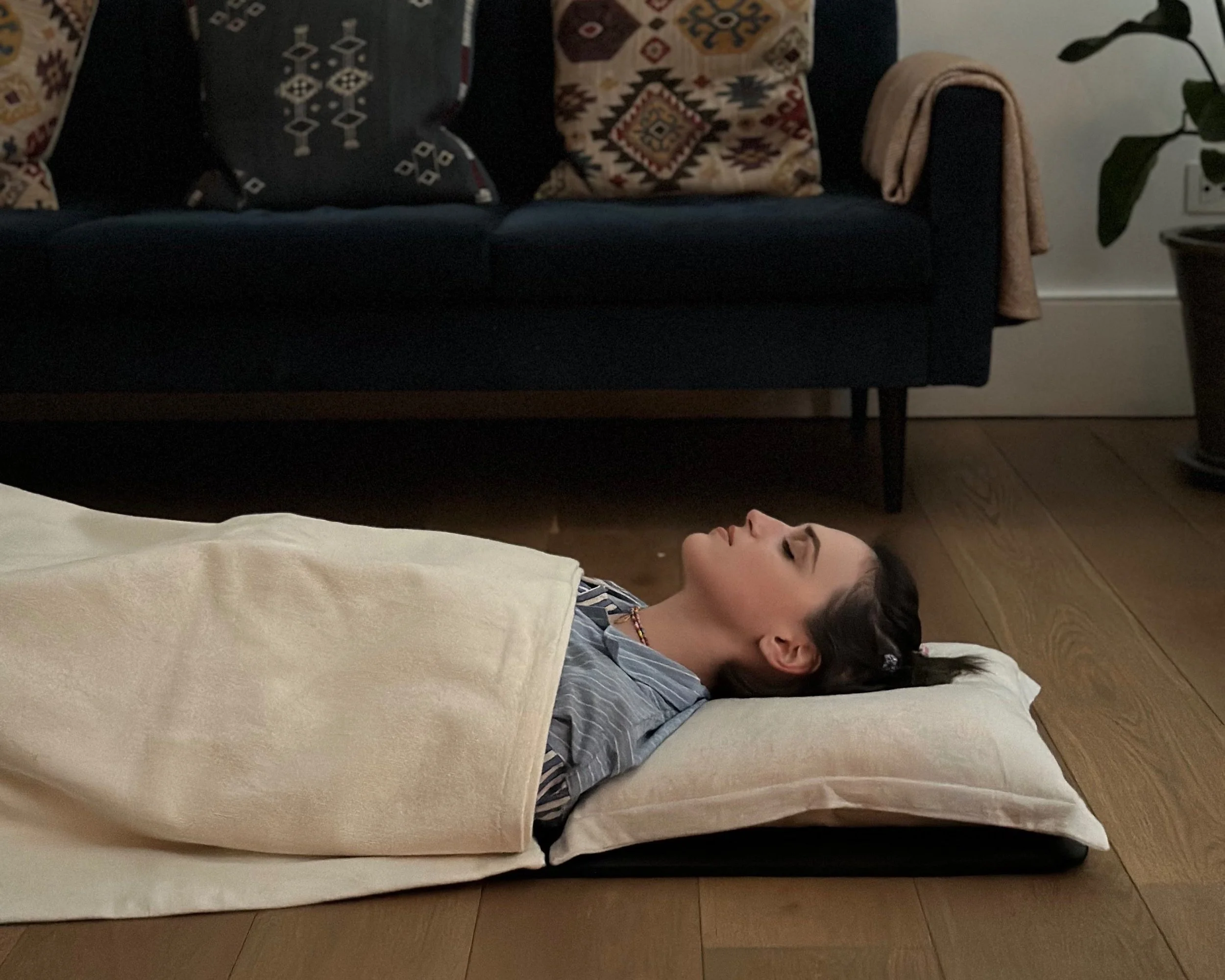A person lying on a pillow and mat on a wooden floor, covered with a beige blanket. In the background, there is a dark sofa with decorative pillows and plants near it.