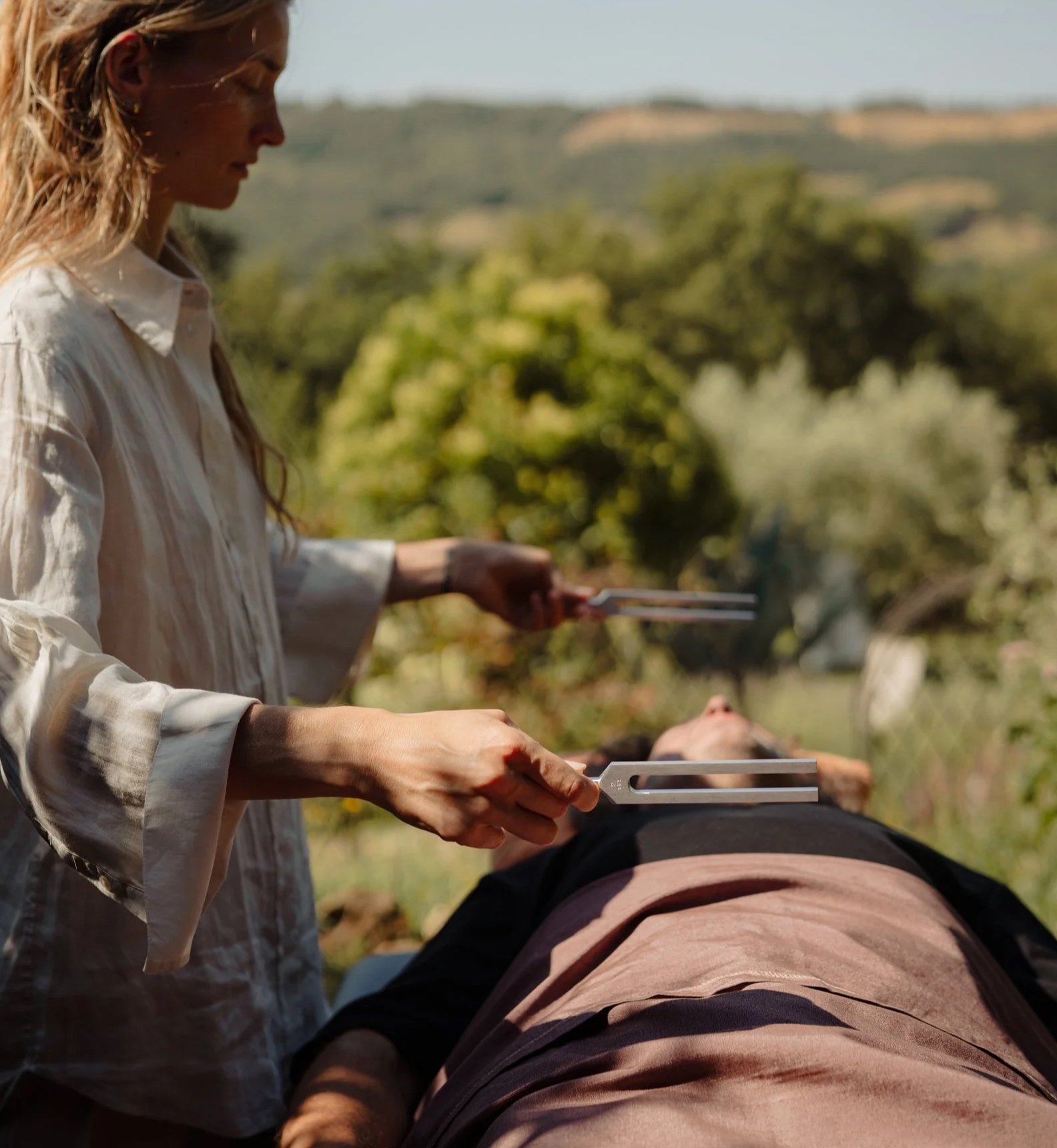 Woman holding tuning forks near her face, sitting in a room with a plant and white shutters.