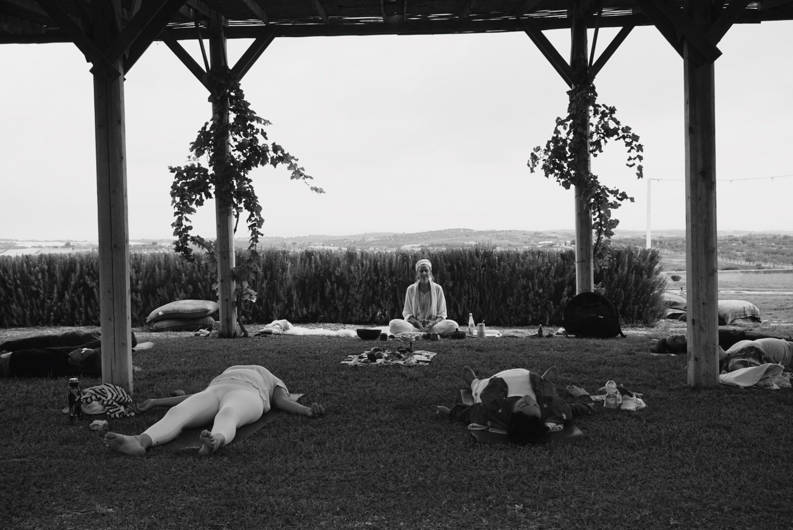 People practicing yoga outdoors under a wooden pergola, with one person meditating and others lying on the grass.