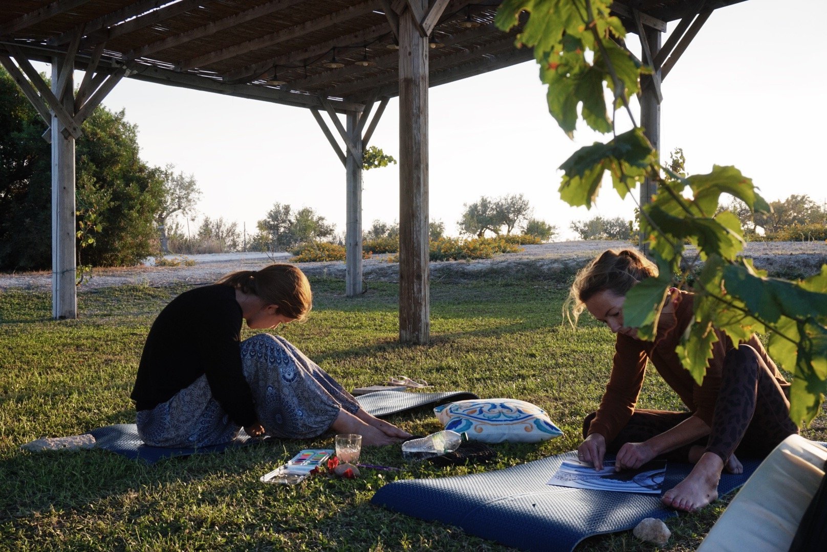 Two people sitting on yoga mats under a wooden pergola outdoors, engaged in drawing or crafting activities on the grass.