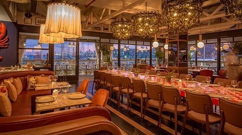 Indoor restaurant with wooden furniture, table settings, and large potted cactus plant, overlooking an outdoor patio with umbrellas and a view of the water and city skyline in the background.