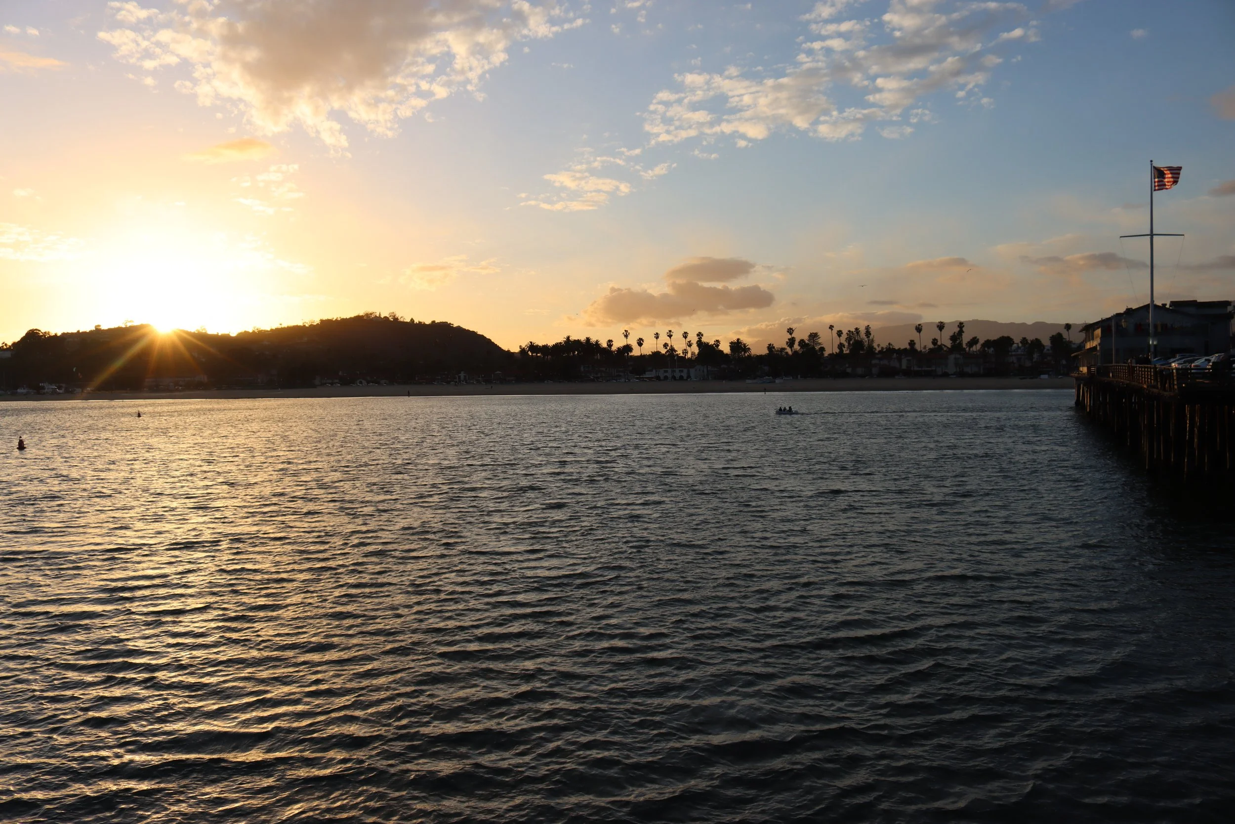 Sunset over a harbor with water ripples, a hill in the background, palm trees along the shoreline, a boat on the water, and a pier with an American flag.
