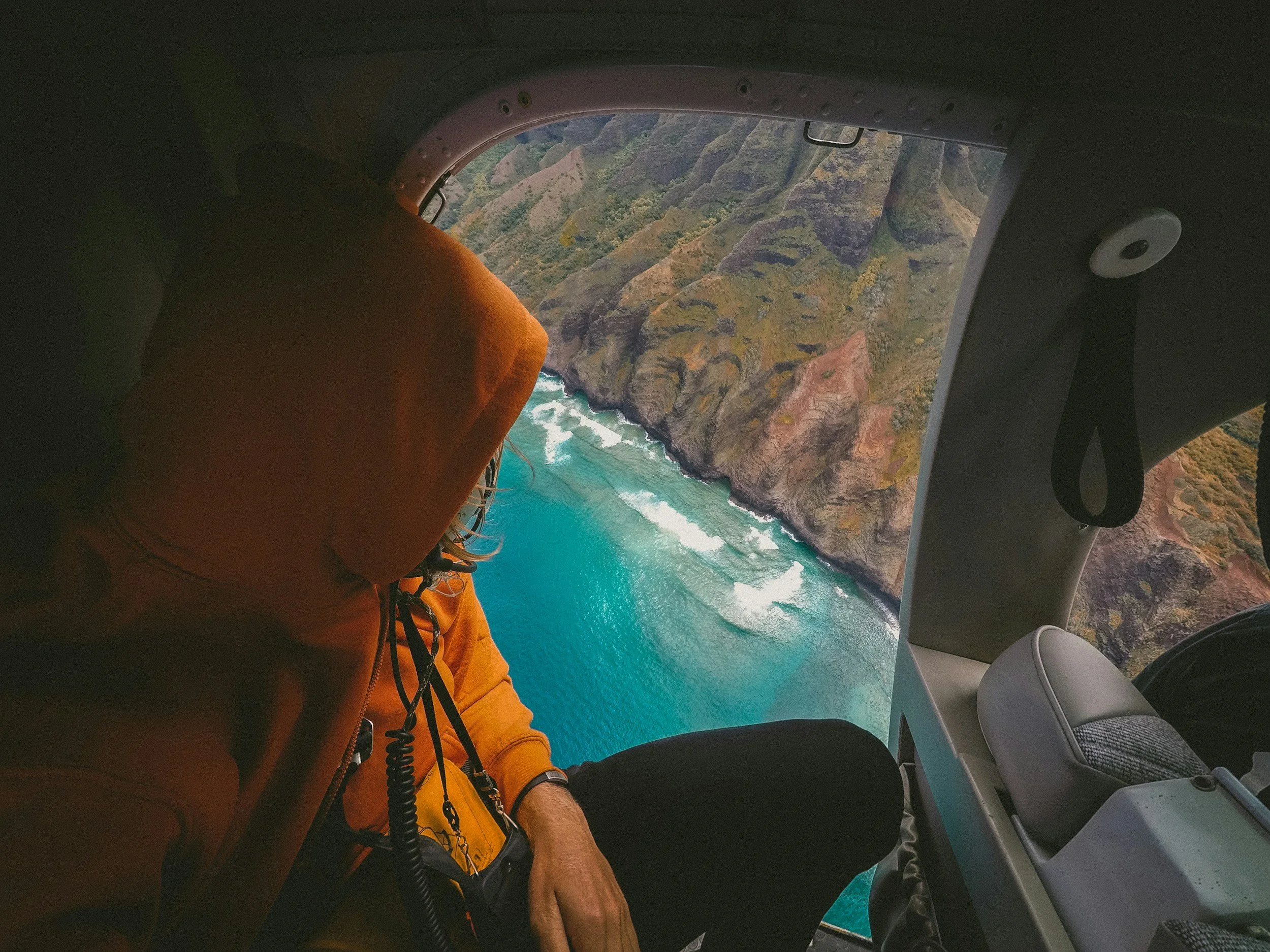 Aerial view from a helicopter over a coastline with turquoise water, cliffs, and lush green hills.