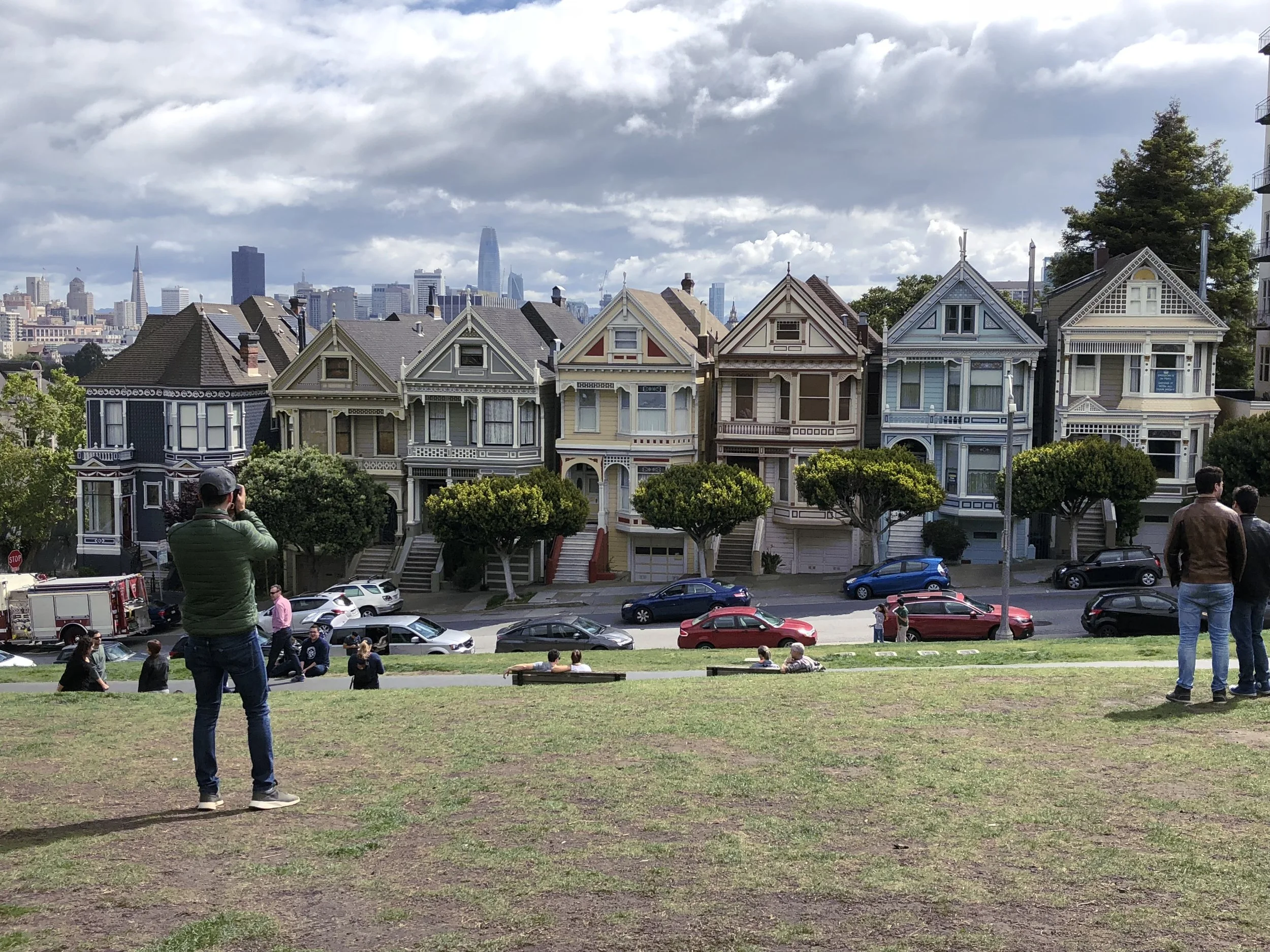 View of the Painted Ladies Victorian houses in San Francisco, with a city skyline and cloudy sky in the background, and people sitting and standing on a grassy hillside.