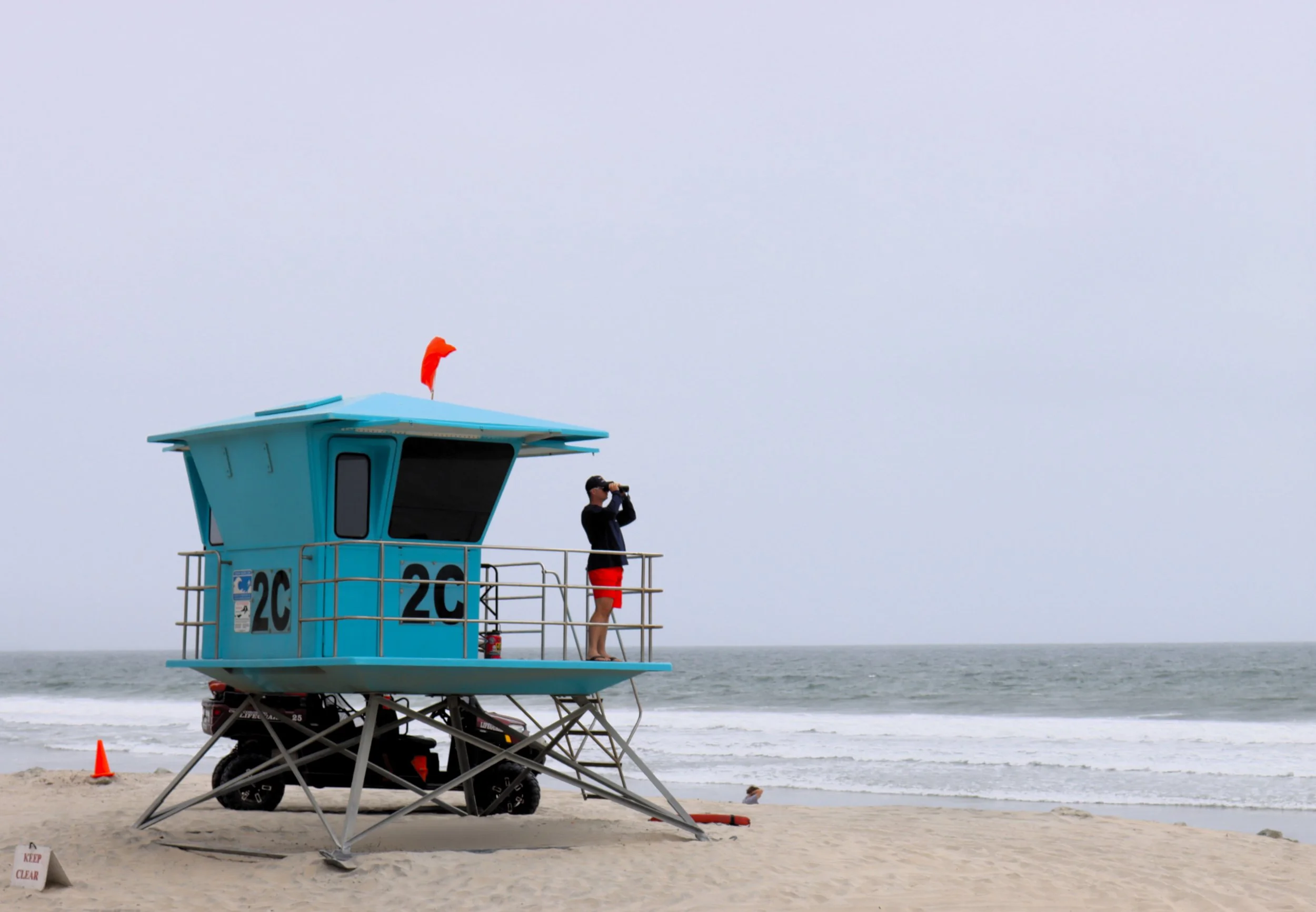 A lifeguard tower on the beach with a lifeguard using binoculars, overlooking the ocean on an overcast day.
