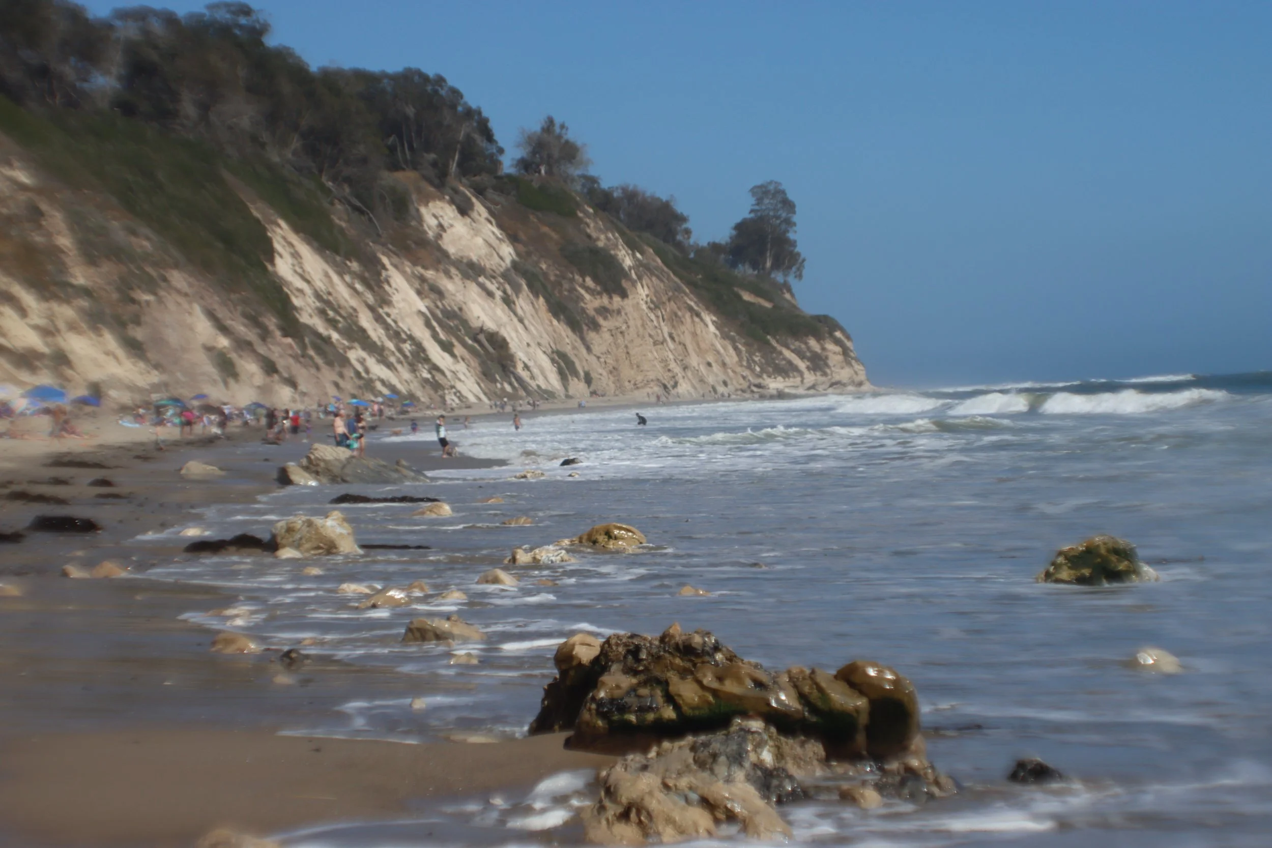 A sandy beach with rocks along the shore, ocean waves, a cliff with trees in the background, and people under umbrellas